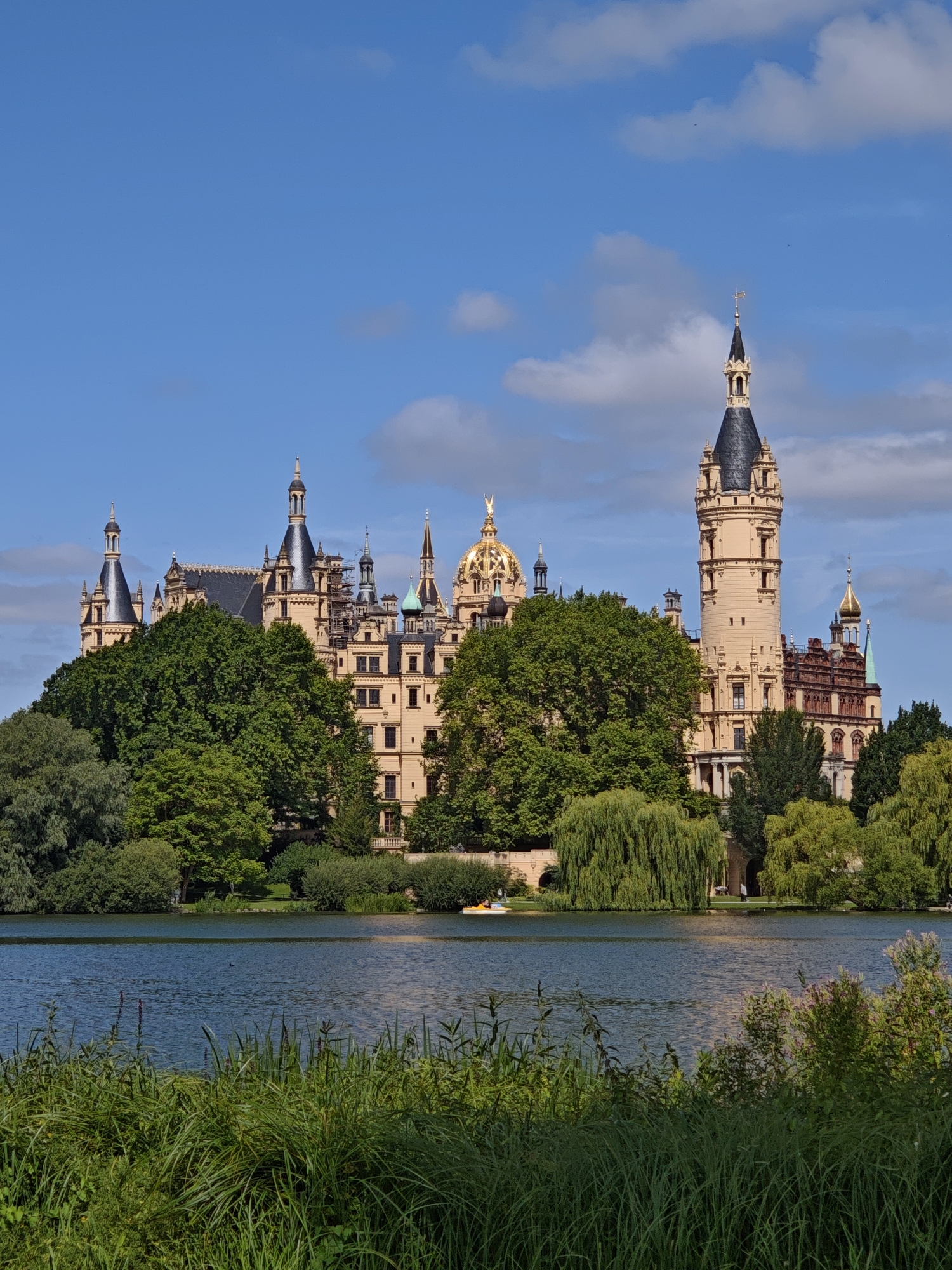 The image depicts a scenic view of Schwerin Castle, a magnificent palace located on an island in the city of Schwerin, Germany. The castle, with its distinctive towers, domes, and elaborate architecture, is set against a backdrop of a clear blue sky dotted with white clouds. Lush green trees surround the castle, partially obscuring its lower levels and creating a picturesque contrast with the pale yellow facade of the building.
In the foreground, a tranquil body of water, likely a lake, reflects the sky and the surrounding greenery. Tall grasses and reeds grow along the water's edge, adding a touch of natural beauty to the scene. A small yellow paddleboat is visible on the water in the middle ground, suggesting recreational activity. The overall impression is one of serene beauty and historical grandeur.