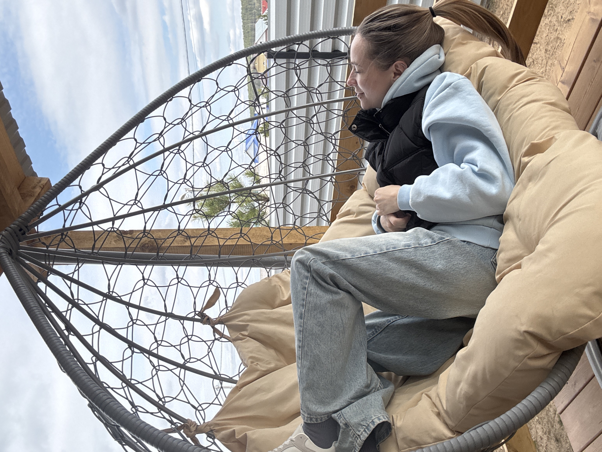 A woman is sitting in a hanging chair, likely outdoors. She has light brown hair pulled back in a ponytail and is wearing a light blue sweatshirt, a black vest, and light-wash jeans. Her left foot is visible and she is wearing white sneakers with dark socks. The hanging chair is made of dark gray metal with a woven design and has a large beige cushion. The chair appears to be on a deck or platform, with wooden planks visible underneath. Through the woven metal, a landscape can be seen, including bodies of water, trees, and buildings. The location is in the city of Chaikovsky, Russia. The woman is looking out at the view.
