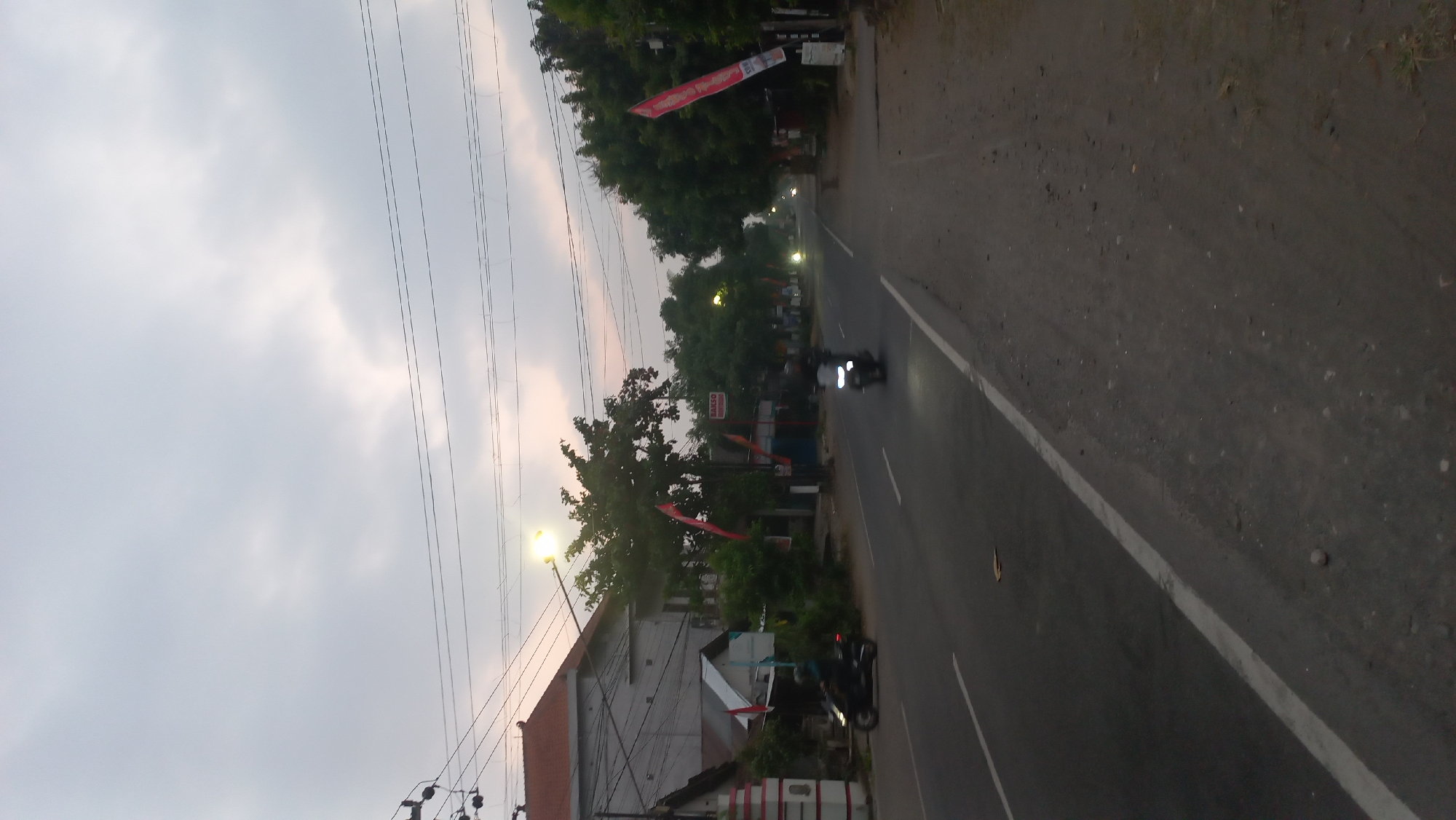The image captures a street scene in Pare, Indonesia, during what appears to be early morning or dusk. The sky is overcast with soft, diffused light. A paved road runs through the center of the frame, with a white line marking the lane division. On the left side of the road, a building with a tiled roof and what looks like a white and red striped pillar is visible, along with trees and some smaller structures. Several red flags are displayed on poles along the roadside.
On the road, two motorcycles are present. One is moving in the left lane and is predominantly black. Another motorcycle is further down the road in the right lane, with its headlight illuminated. There are also several streetlights visible, some of which are lit, casting a warm glow. The right side of the road is unpaved, with dirt, small stones, and some sparse vegetation. Overhead, power lines are strung across the scene, supported by poles. A sign that says "BAKSO" is visible on the left side. The overall atmosphere is calm and somewhat subdued due to the lighting conditions.