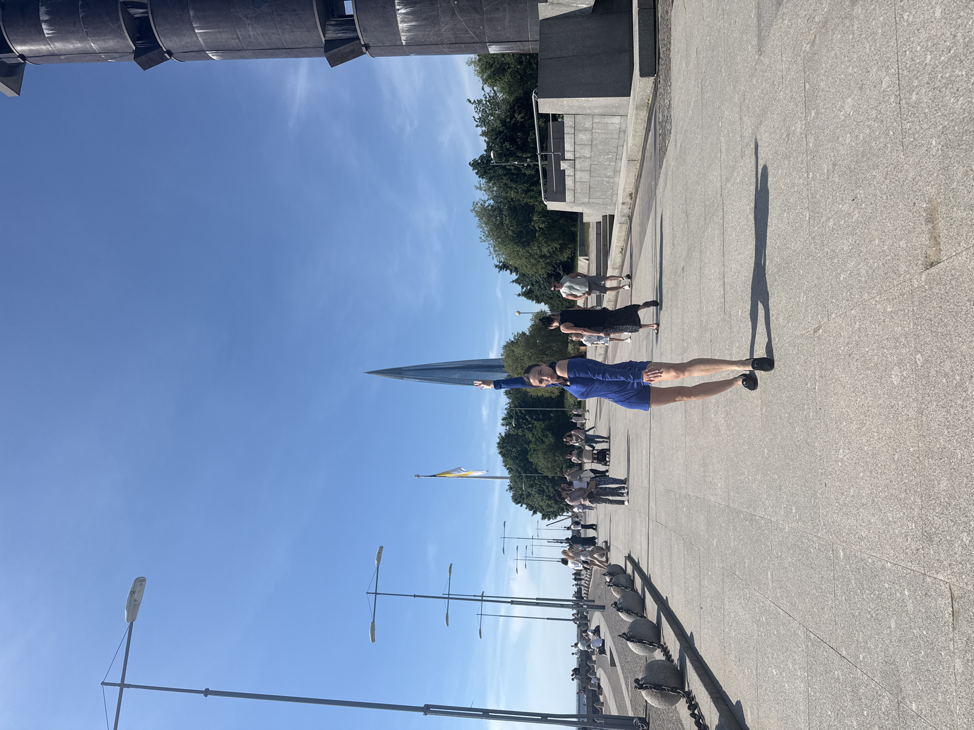 A woman in a blue off-the-shoulder dress stands on a paved promenade in Saint Petersburg, Russia, with her right arm raised. The promenade has a seawall with large, round, chained buoys along the left side, and several lampposts line the walkway. In the background, a tall, slender, modern skyscraper with a pointed top dominates the skyline, identified as the Lakhta Center. To the right of the skyscraper, a large, dark, cylindrical structure, possibly a monument or part of an industrial facility, is visible. A Russian flag, yellow and white, flies from a tall flagpole between the skyscraper and the dark structure. Numerous people are walking on the promenade, some with strollers, and others are seated along the seawall. The sky is clear and blue with a few wispy clouds. The scene appears to be a sunny day with a bright atmosphere.