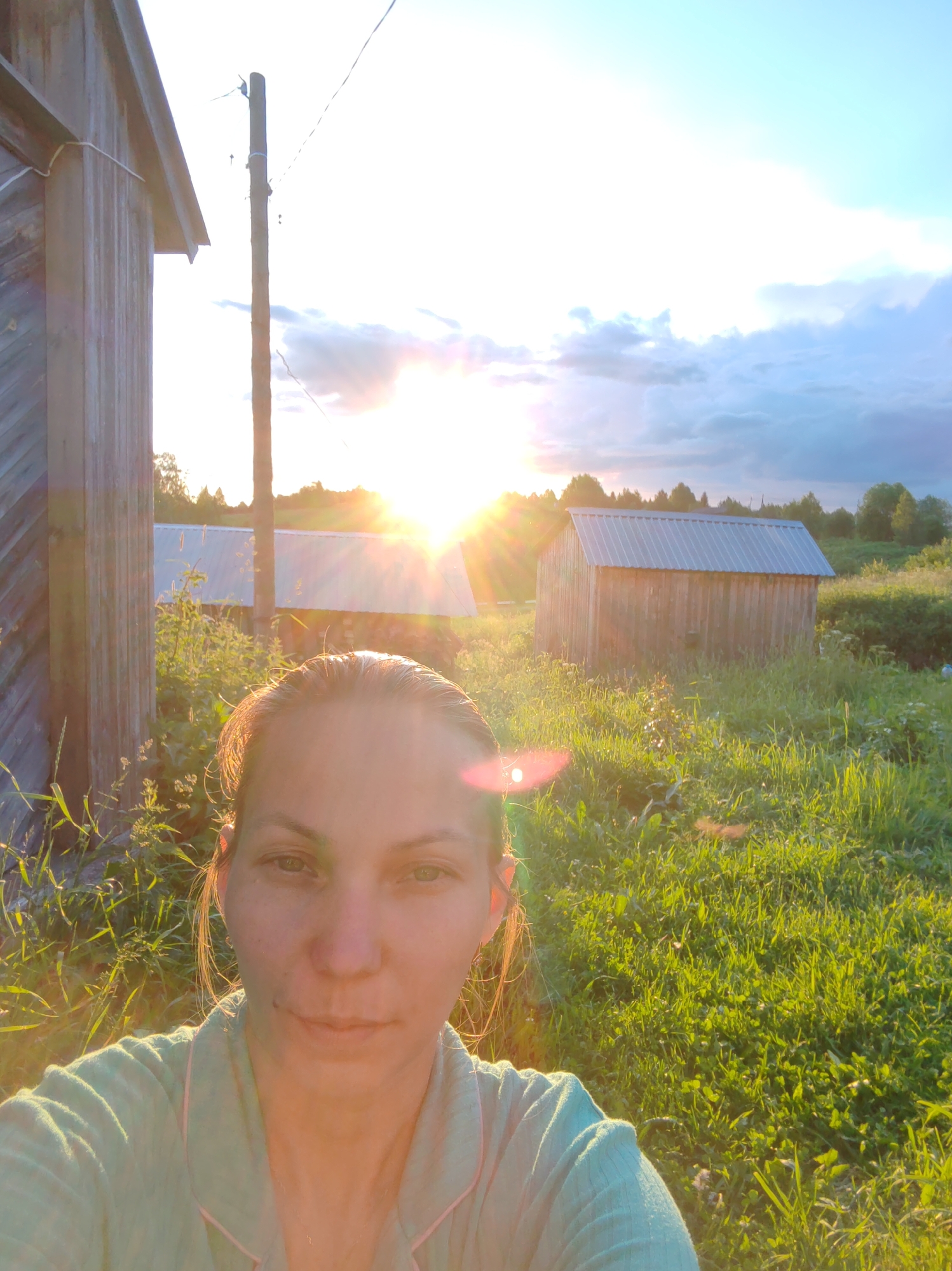 A selfie is taken outdoors at sunset in a rural environment. The foreground features the face of a woman looking directly at the camera. She has light skin, light brown hair, and is wearing a light blue collared shirt. Behind her, the sun is low in the sky, casting a warm golden light across the scene and creating lens flares. To the left of the frame, a wooden structure, possibly a shed or outbuilding, is partially visible. A wooden utility pole stands between the woman and the setting sun. Further in the background, another wooden shed with a corrugated metal roof is visible amidst tall green grass and vegetation. The sky above is a mix of blue and clouds, with the sun’s rays creating a bright halo effect. The overall setting appears to be a countryside or village in Чухлэм, Russia.