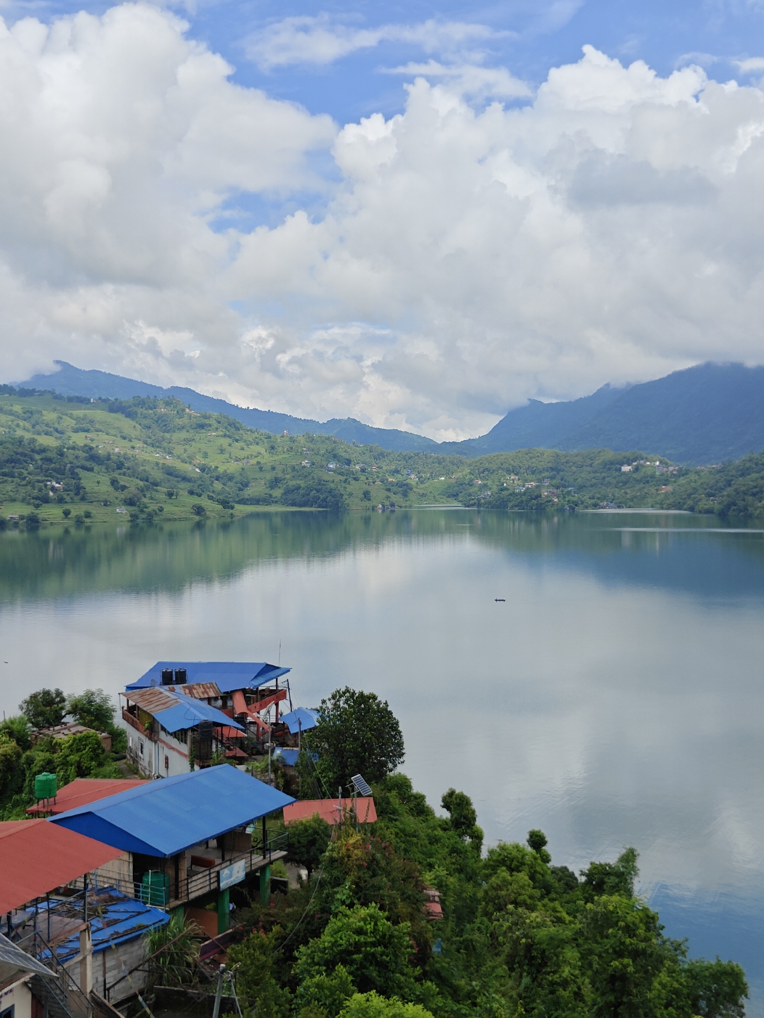 The image depicts a serene panoramic view of Phewa Lake in Pokhara, Nepal. The foreground is dominated by a cluster of buildings with brightly colored roofs, primarily red and blue, perched on a lush green hillside. These structures appear to be a mix of guesthouses or restaurants, with multiple levels and balconies. Dense foliage, including trees and shrubs, surrounds these buildings.
The middle ground features the expansive, tranquil waters of Phewa Lake, reflecting the sky and the surrounding hills. A small, dark boat with two occupants can be seen in the distance on the lake.
The background showcases rolling, verdant hills that gradually ascend into mist-shrouded mountains under a partially cloudy sky. The clouds are a mix of white cumulus and darker, grayer formations, suggesting a humid or overcast day. The overall atmosphere is peaceful and picturesque, characteristic of the natural beauty of Pokhara.