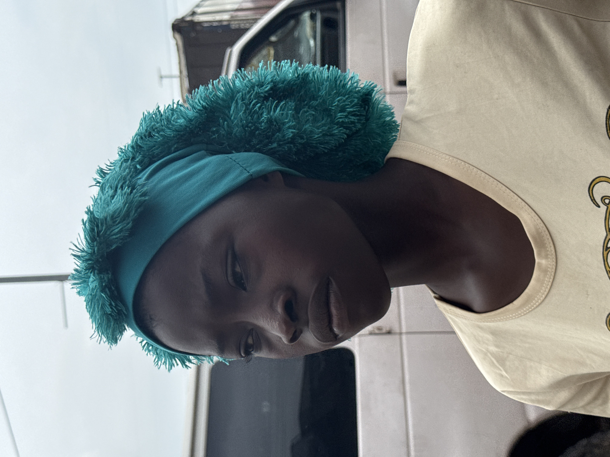 In Lekki, Nigeria, a young Black girl is captured in a close-up, profile shot. She is looking to her left, with her face angled slightly down. The girl is wearing a vibrant teal headwrap adorned with fringe, and a pale yellow t-shirt. Behind her, a white van is visible, with its rear end slightly out of frame. Further in the background, a dark truck and a hazy sky complete the urban environment. The overall lighting suggests an overcast day.