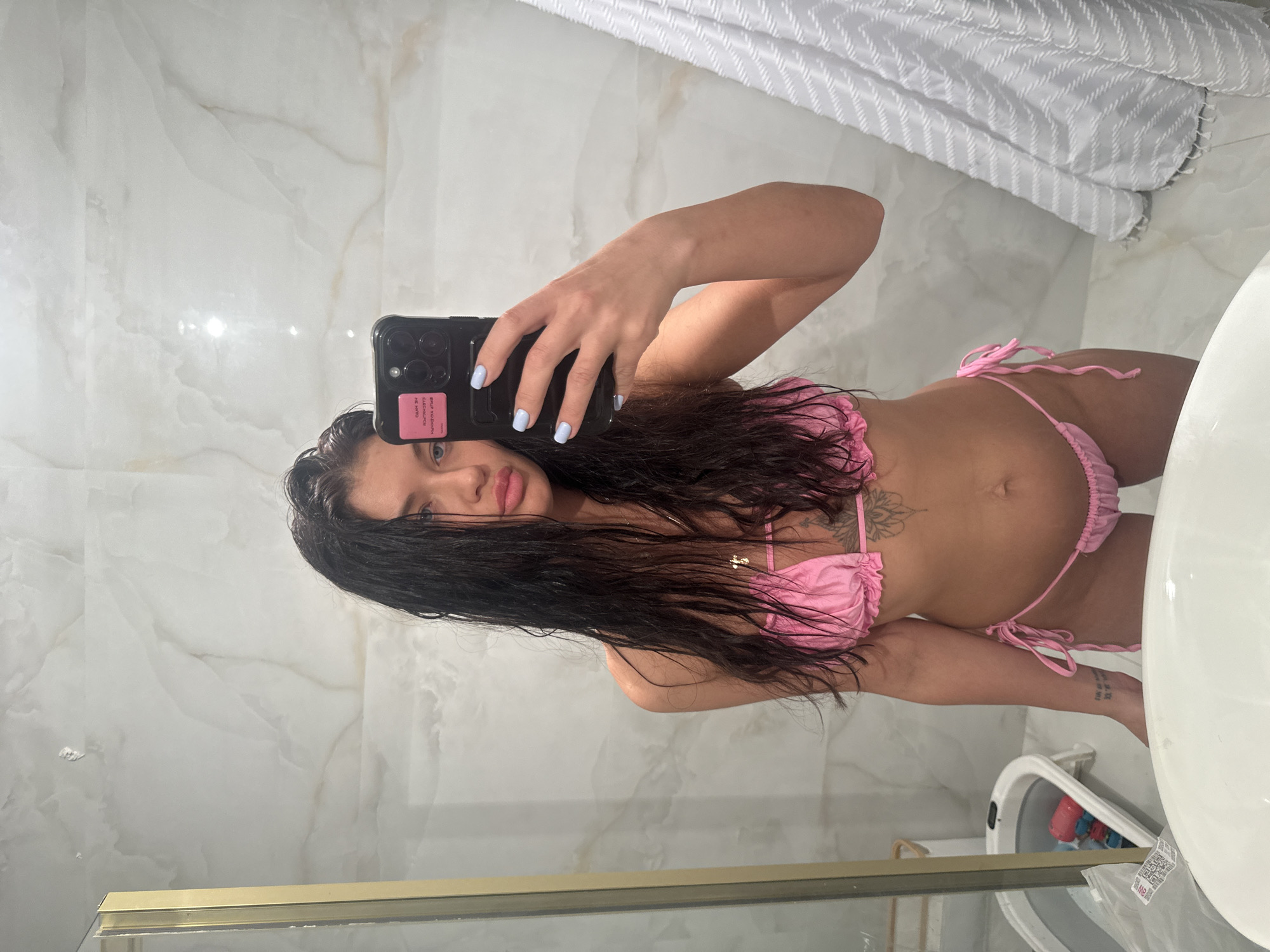 A woman is taking a selfie in a bathroom. She has long, dark, wet hair and is wearing a pink bikini. The bathroom has marble-like wall tiles, a white sink, and a glass shower door. A white, patterned shower curtain is partially visible. There is no indication of the location being Moscow, Russia.
