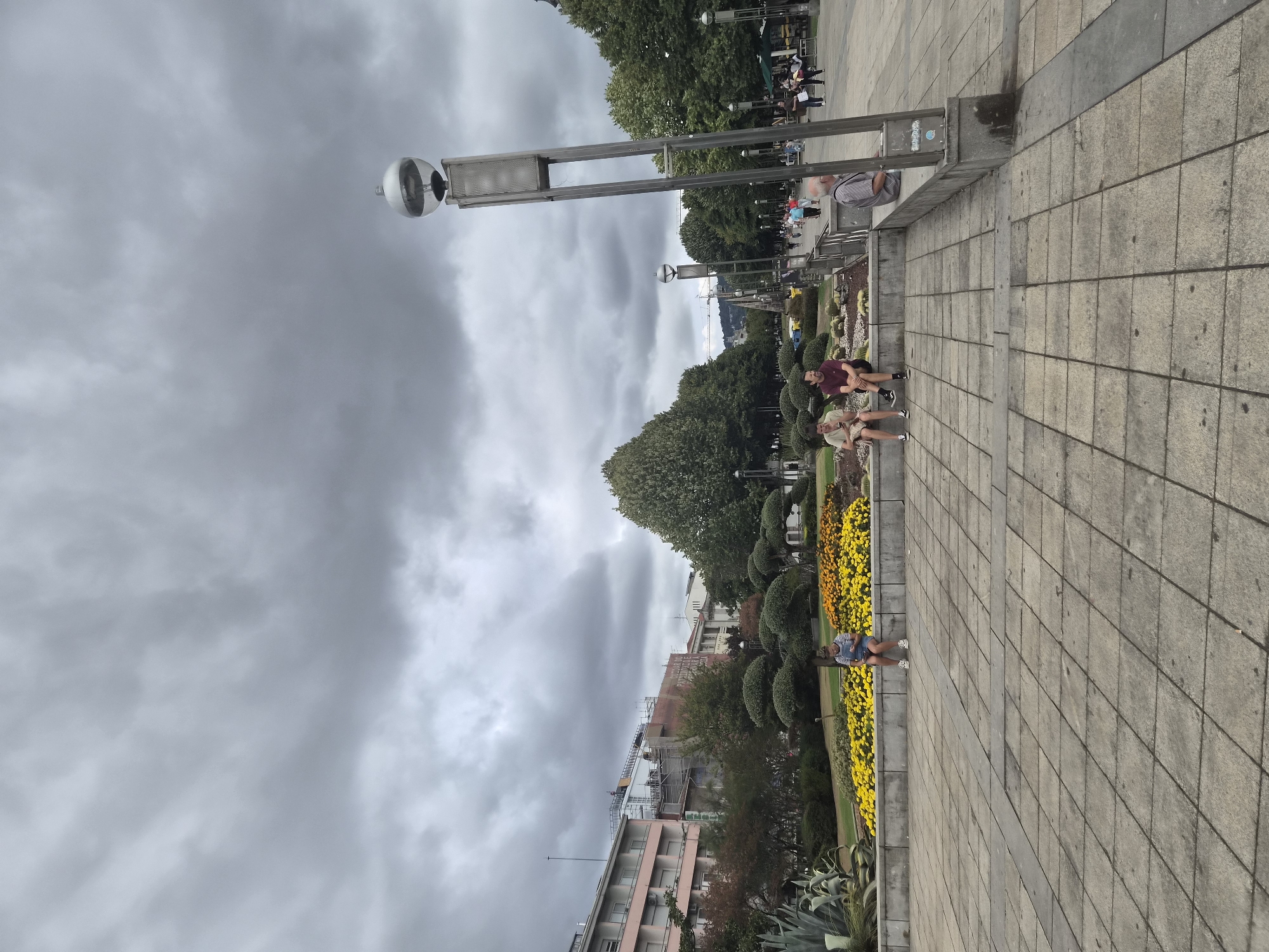The image shows a public square in Braga, Portugal, under a cloudy sky. The foreground is a paved area with large square tiles. Behind the paving is a raised flowerbed filled with yellow and orange flowers. Several people are seated on a low wall bordering the flowerbed. To the left of the flowerbed, there are some large succulent plants. In the background, there are trees, buildings, and streetlights. A young woman in a striped shirt and denim shorts is sitting on the wall, looking at her phone. Two men are sitting further down the wall. Another man is sitting on a bench to the right of the wall, facing away from the camera. In the distance, people are walking along the square, and there are several lamp posts. One prominent lamp post with a spherical light fixture is in the right foreground. A building with a pink facade and several windows is visible in the background to the left. Construction work appears to be underway on a building further back.