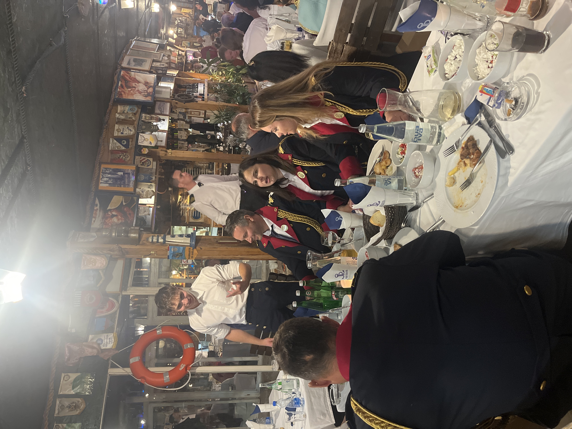 The image captures a lively scene inside a restaurant in Budva, Montenegro. Patrons, some wearing navy blue uniforms with red trim and gold accents, are seated at a long table covered with a white tablecloth. The table is laden with plates of partially eaten food, water bottles, and various glasses. A waiter in a white shirt and dark pants interacts with the diners. The restaurant's interior is decorated with nautical-themed items, including framed pictures and a life preserver, displayed on the walls. The atmosphere appears festive.