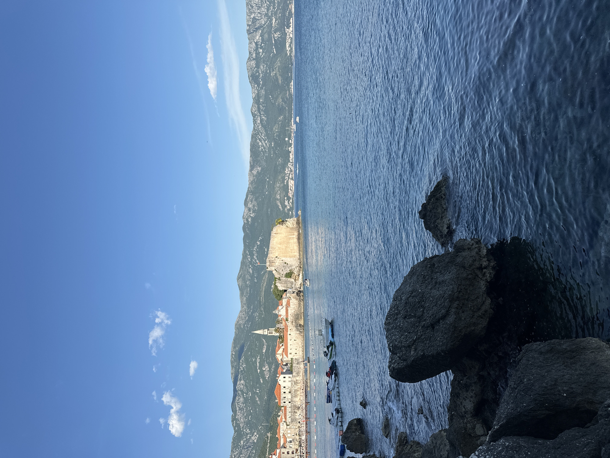 This image shows a coastline view of the city of Budva, Montenegro. The sea occupies a significant portion of the right side of the frame, with dark, rocky outcrops along the shoreline in the foreground. The old town of Budva is visible along the coast, characterized by its stone buildings, red-tiled roofs, and a prominent church tower. A medieval fortress is perched atop a hill above the old town. Mountains rise in the background, framing the cityscape. The sky is blue with sparse white clouds. Several small boats are moored near the shore.