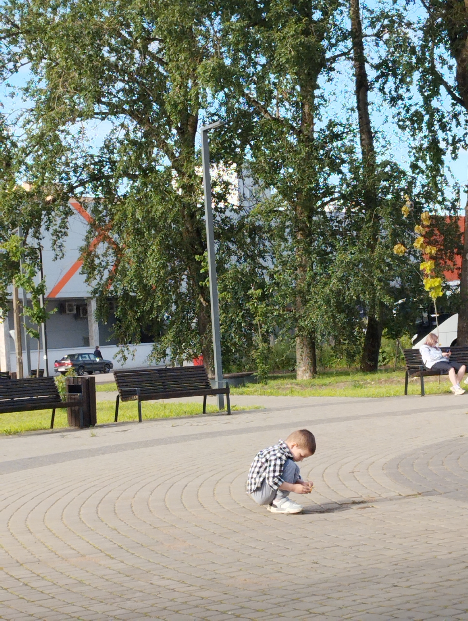 In Kingisepp, Russia, a young boy wearing a plaid shirt and grey pants crouches on a patterned brick walkway. He is intently examining a small object in his hands. To his left, a dark trash receptacle stands next to a dark slatted bench. Further to the left, another identical bench is partially visible. In the background, a dark-colored car is parked, and a person is standing near it. Beyond the car, a building with a white facade and an orange diagonal stripe is visible, along with air conditioning units. The scene is framed by lush green trees, and the sky is a clear blue. To the right of the boy, further down the walkway and partially obscured by trees, another dark slatted bench is occupied by a woman sitting and looking at her phone. The overall setting appears to be an outdoor public space with an urban backdrop.