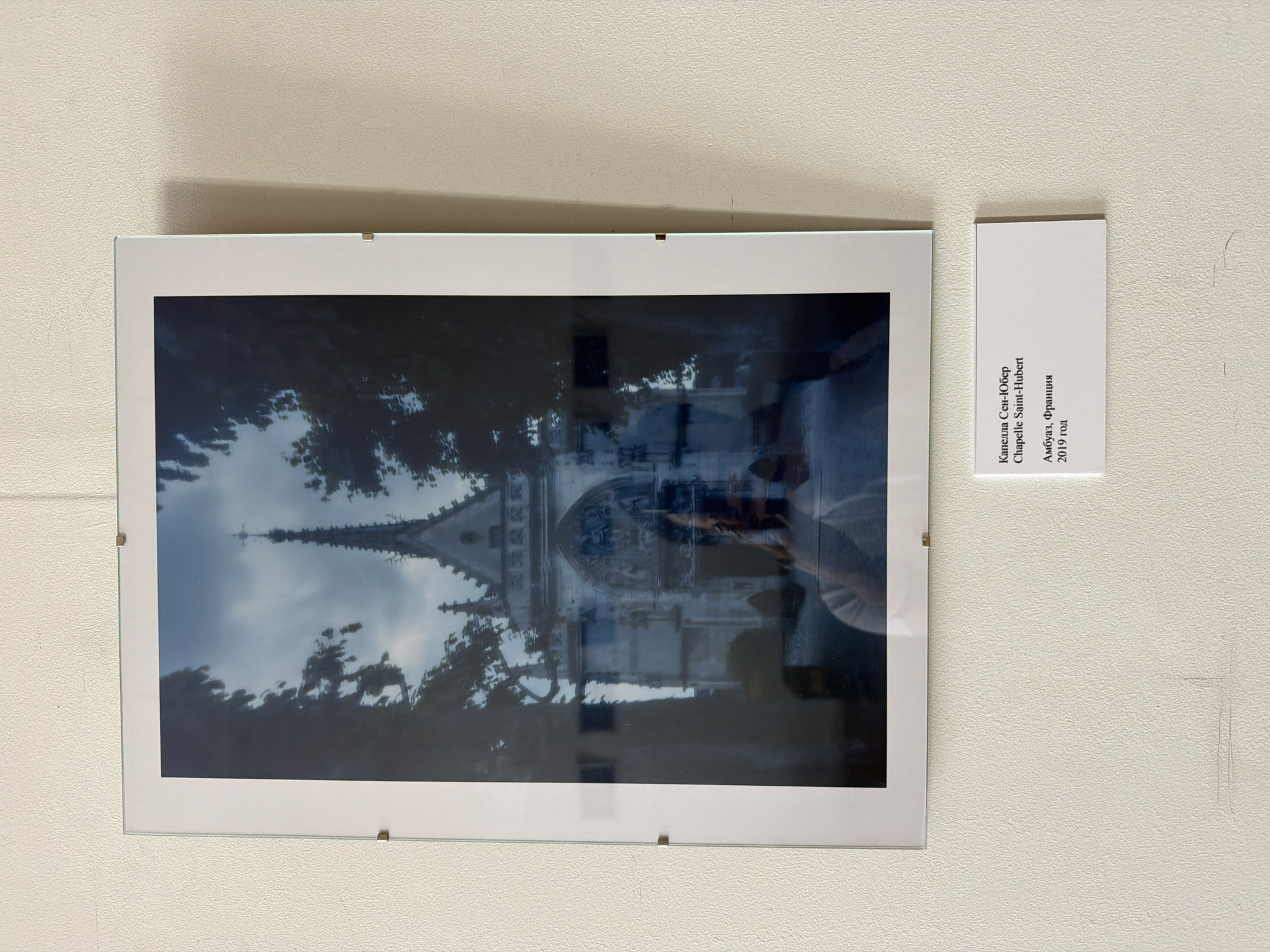 A framed photograph is displayed on a textured, off-white wall. The photograph depicts a Gothic-style building with a spire, partially obscured by dark trees on either side. A reflection in the glass of the frame shows a person holding a camera, taking the photograph. Below the framed photograph, a small white label is affixed to the wall. The label contains text in Russian and French, indicating the subject is "Chapelle Saint-Hubert" in "Amboise, France" from "2019". The wall appears to be a gallery or exhibition space, with a thin wire visible near the top of the frame, suggesting a hanging mechanism. The overall scene is well-lit, with the focus on the artwork. There is no visible indication of the location being Nizhny Novgorod, Russia; the label clearly states the location is Amboise, France.