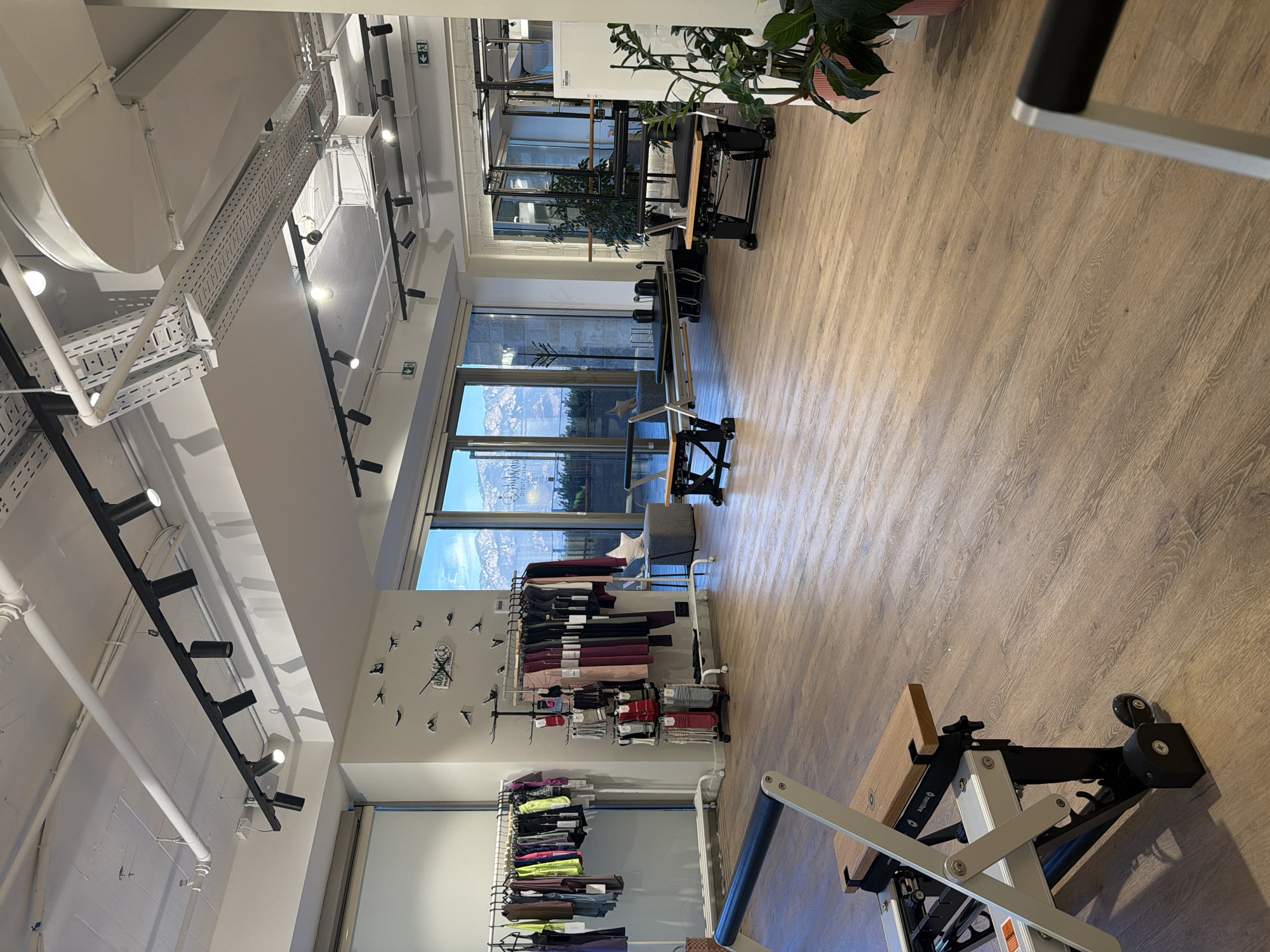 The image depicts the interior of a modern, well-lit pilates studio in Boreti, Montenegro. The studio features a polished wooden floor and high ceilings with exposed ductwork and a track lighting system. Two pilates reformers, with wooden and metal components, are positioned in the foreground and mid-ground. Racks of athletic apparel, including leggings and tops, are displayed on the left and in the mid-ground, with a rack of socks to the right of the apparel. A wall clock with a whimsical bird design adorns the wall in the mid-ground. Large windows in the background offer a view of mountains and what appears to be the Adriatic Sea. To the right, a large plant in a pink pot adds a touch of greenery. The space appears clean, organized, and ready for use. There are no people visible in the image.
