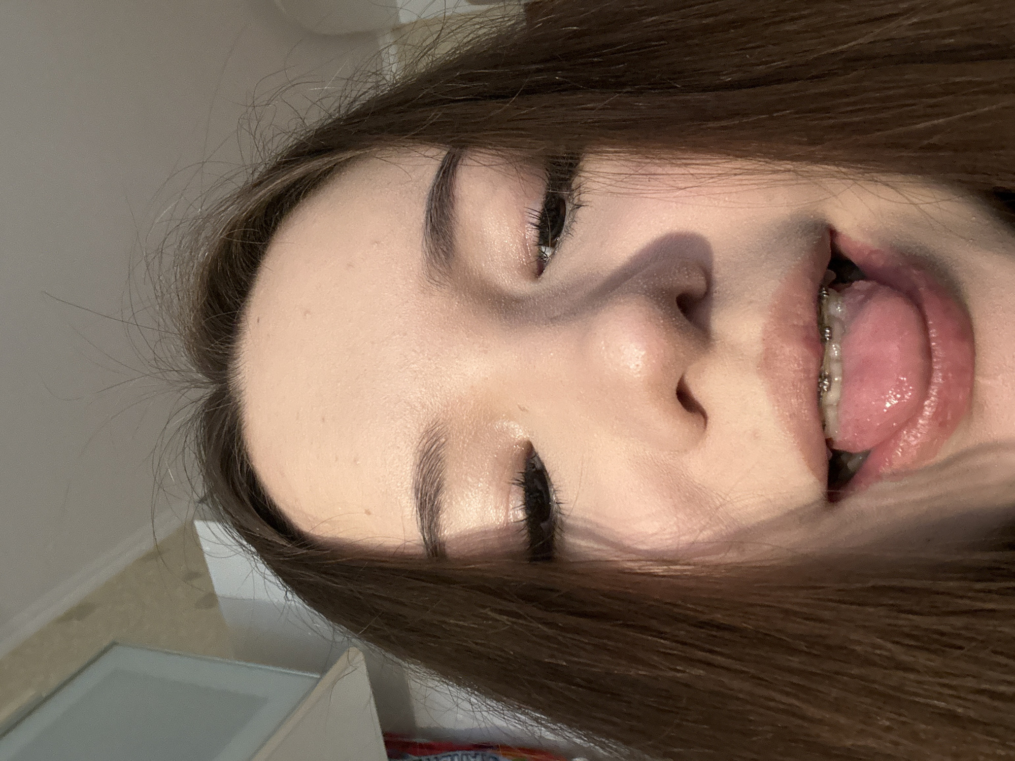A close-up, sideways portrait captures a young person, likely female, with long, dark brown hair, positioned against a light, out-of-focus background. The image is rotated, presenting the individual's face with their head tilted. The subject is making a playful expression, sticking out their pink, wet tongue. Braces are clearly visible on their upper teeth. One eye is partially closed, showing long eyelashes and a well-defined eyebrow, while the other eye is largely obscured by hair. The skin appears clear with a subtle sheen, possibly from natural oils or cosmetic highlighting. The background suggests an indoor setting, with blurred light-colored walls or a ceiling, and faint indications of architectural trim. No text is visible in the image. The overall impression is one of lightheartedness and a candid moment.