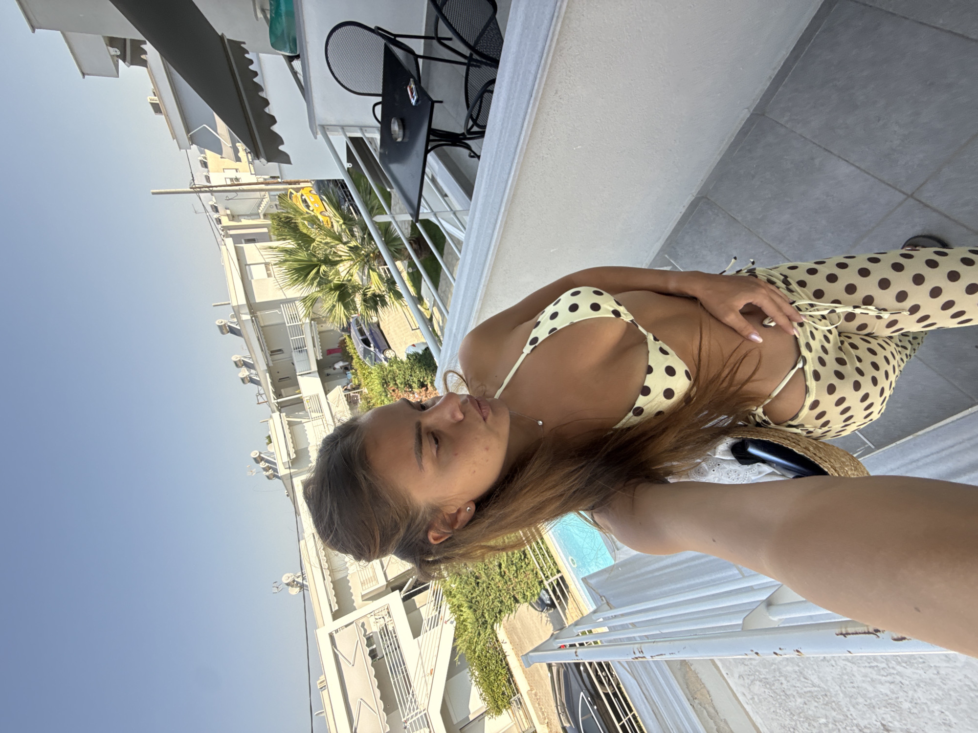 A woman stands on a balcony in Ofrynio Beach, Greece, wearing a beige bikini with brown polka dots and matching patterned pants. She holds a straw bag in her left hand and her right hand is on her hip. Behind her, a swimming pool is visible, along with buildings, palm trees, and parked cars. The sky is clear and blue.