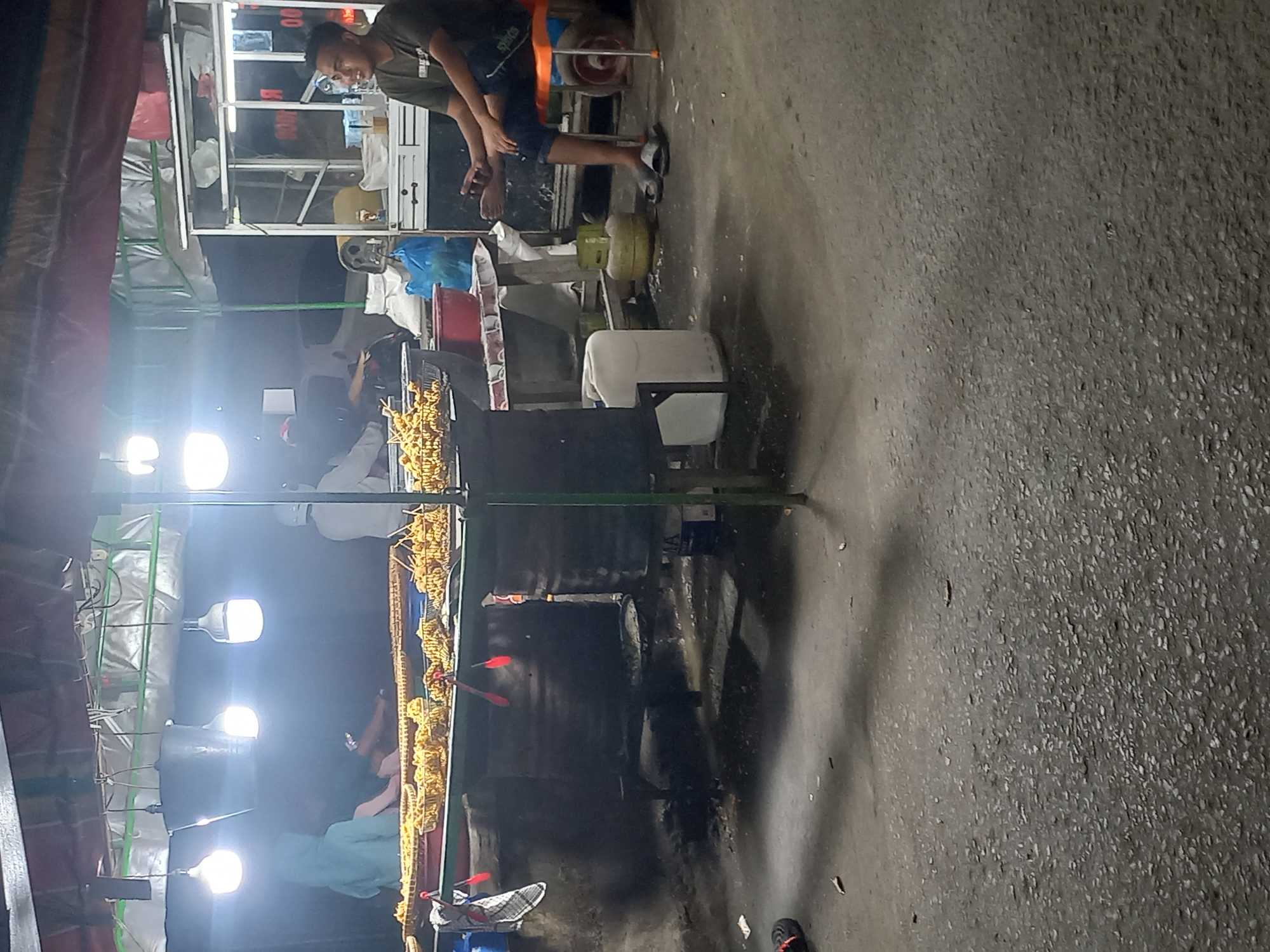 This image depicts a vibrant night street food scene in Idi Rayeuk, Indonesia, set along a paved road. The time of day is clearly night, as indicated by the bright artificial overhead lights illuminating the stalls and their surroundings against a dark background.
The primary subjects are people engaged in the preparation and selling of street food. A young male vendor, wearing a dark green t-shirt with "SPORTS" printed on it and dark shorts, stands prominently at one of the food stalls. He has short, dark hair and appears to be interacting with customers or working at the counter. His feet are visible, one bare and one in a sandal. Further down the line of stalls, other individuals are present, including one person in a light blue shirt.
The scene is dominated by several interconnected food stalls with simple metal frames and white or translucent coverings. The most visible stall showcases long trays filled with golden-brown, coiled, fried snack items, possibly spiral potatoes or fried noodles, ready for sale. Red serving tongs are placed among these snacks. Adjacent to the display area are large cooking sections, characterized by dark, possibly sooty surfaces, suggesting the use of deep fryers or woks.
Various objects related to the operation of the stalls are scattered around: white and red plastic buckets, and two yellow-green gas cylinders, which likely provide fuel for cooking. Multiple bright white LED or fluorescent lights hang overhead, casting a strong glow and creating a lively atmosphere. The ground is a dark, rough, paved surface. Some blurry, illegible text is visible on a white cabinet or sign behind the main vendor. The scene portrays a typical bustling street food environment common in Southeast Asia at night.