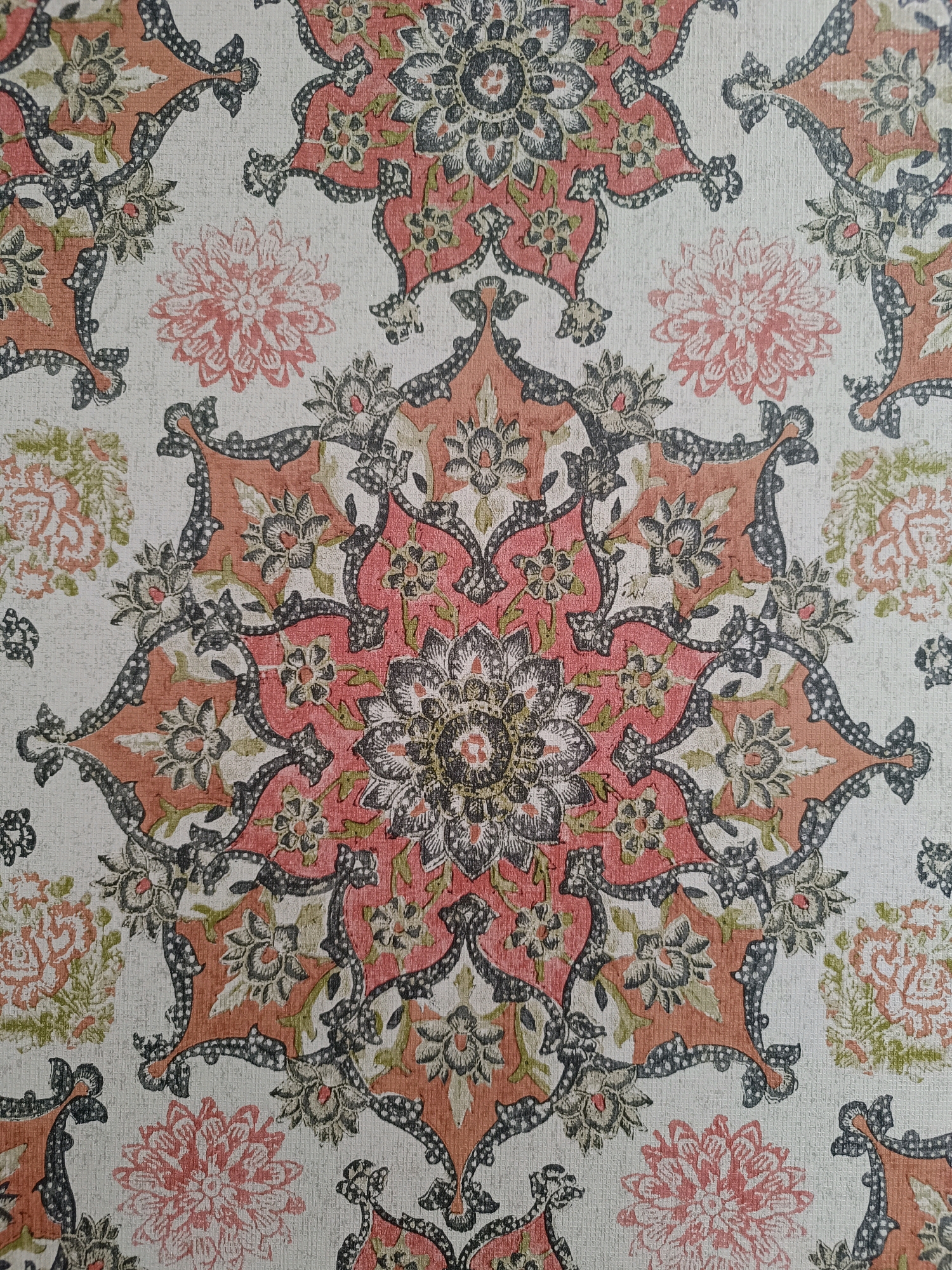 The image shows a close-up of a wallpaper with a floral and paisley-like pattern. The dominant colors are a muted coral or salmon pink, grey, black, and off-white, with hints of muted green. The pattern is intricate and symmetrical, featuring a central star-like motif with floral elements radiating outwards. Smaller floral and leaf designs are interspersed within the larger pattern. The texture of the wallpaper is visible, suggesting it may be a textured or fabric-backed material. The lighting appears to be even, without harsh shadows, indicating an indoor setting or diffuse natural light. There are no people or any discernible elements that would place this image specifically in Srinagar, India.