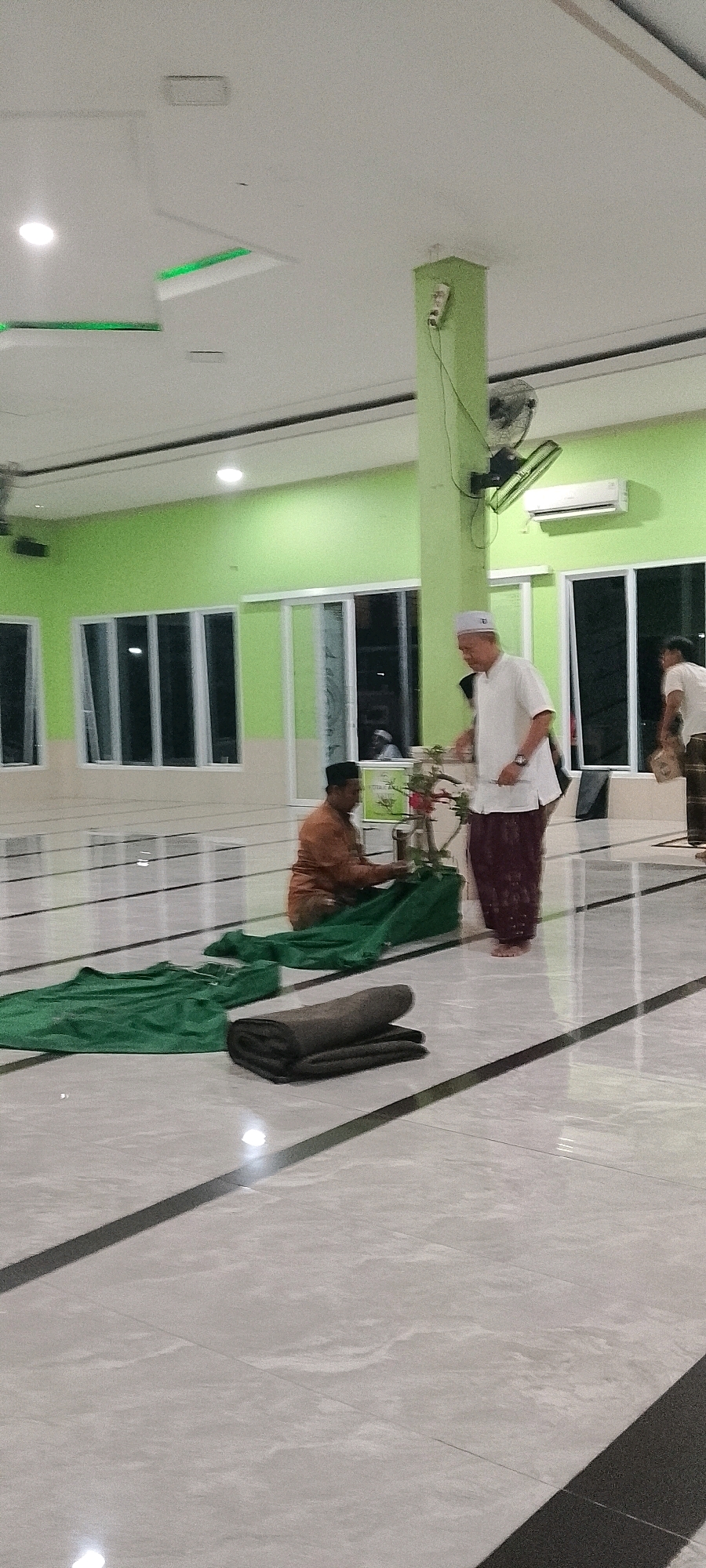 In a brightly lit room with lime green walls and a white ceiling, a man wearing a white cap and shirt, and a dark patterned sarong, stands on a checkered tiled floor. He is interacting with a table that holds a plant with red flowers. In front of him, a large, unfolded green cloth lies on the floor, along with a folded dark blanket. To his left, a man in an orange shirt and a black cap is seated on the floor, also near the green cloth. The room features large windows with white frames and reflective glass, and ceiling fans are visible. A wall-mounted air conditioner is also present. The overall environment appears to be a community hall or a place of worship, possibly in Satriamekar, Indonesia, given the context.