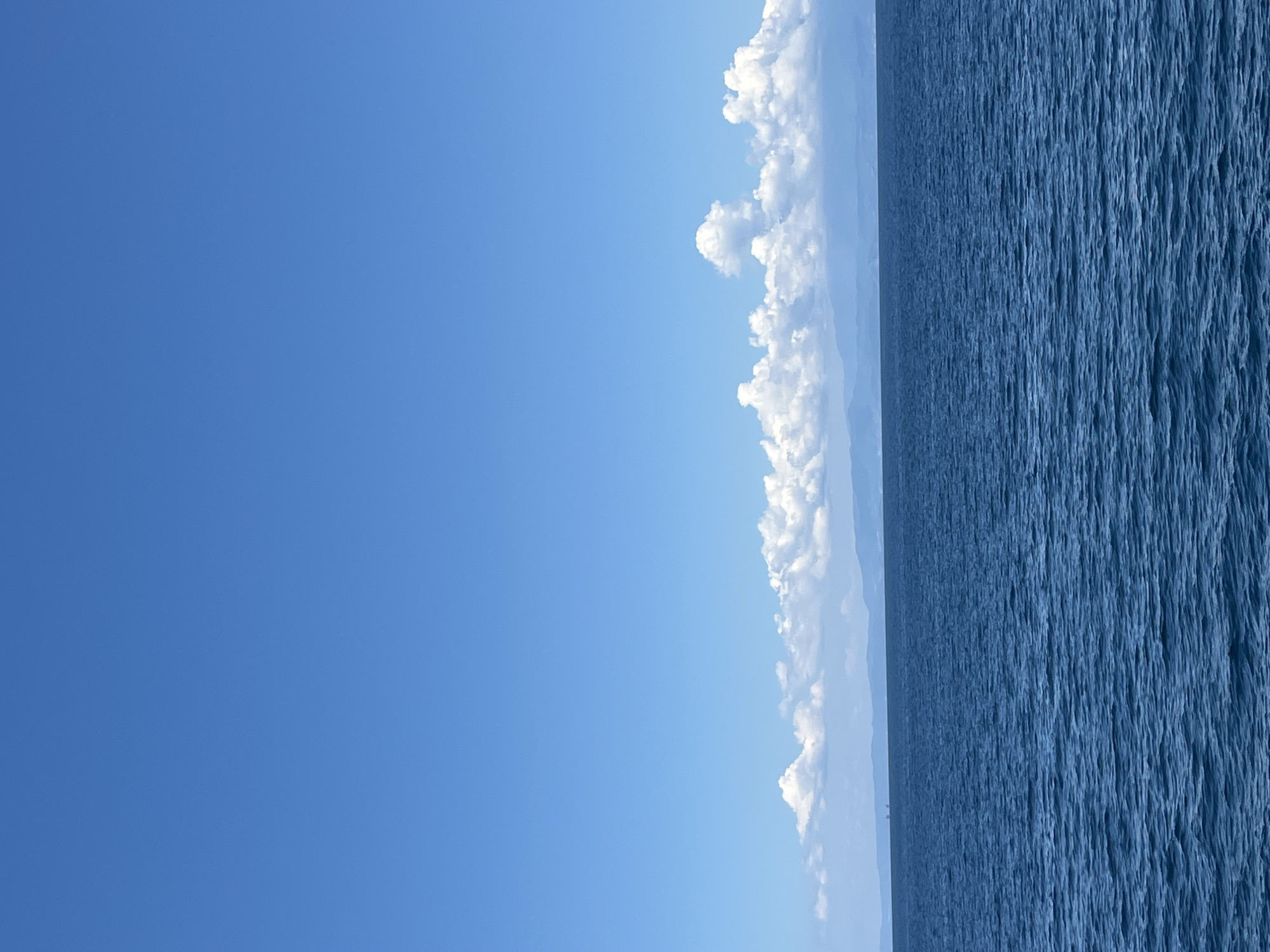 The image depicts a vast expanse of deep blue ocean under a clear, bright blue sky. The water exhibits gentle ripples, creating a textured surface. In the distance, along the horizon, a line of hazy, mountainous land is visible, suggesting the presence of an island. A band of fluffy, white clouds hovers above the landmass, adding depth to the scene. There are no people or animals visible in the image, and no specific structures or landmarks are discernible on the distant land. The overall impression is one of a serene and open seascape. While the specific location within Thassos, Greece cannot be determined from the image alone, the scene is consistent with the coastal geography of the island, which is known for its beautiful beaches and clear waters.