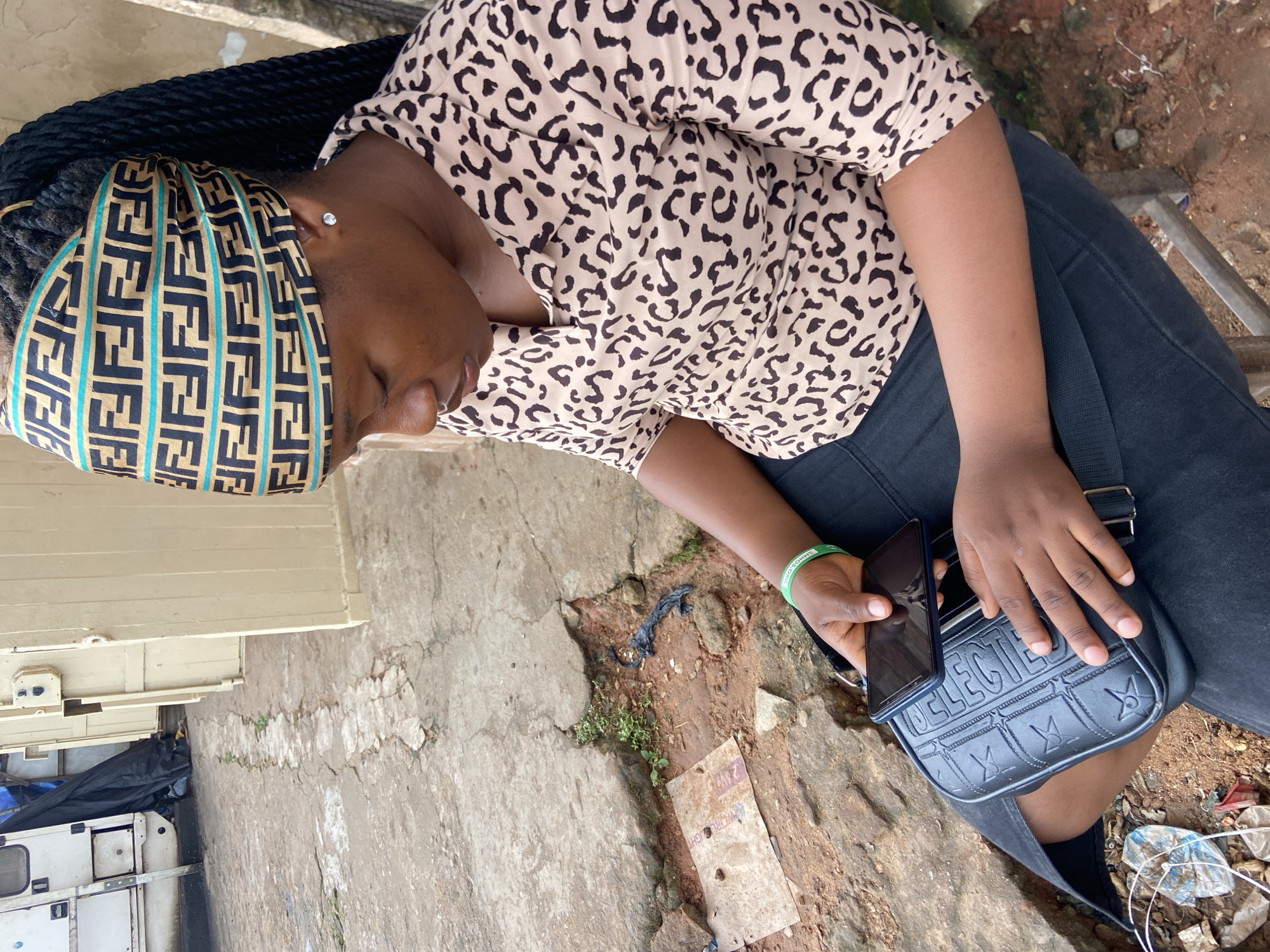 In Nkpor, Nigeria, a young woman sits outdoors on a stool, her gaze directed downwards at her smartphone. She wears a leopard-print blouse and dark pants, accessorized with a patterned headscarf and a green bracelet. A black crossbody bag rests on her lap, and her hands are engaged with the phone. The surrounding environment is a rough, cracked concrete ground with scattered debris. In the background, a white vehicle and light-colored doors are visible. The scene suggests an informal, possibly public space within the city.