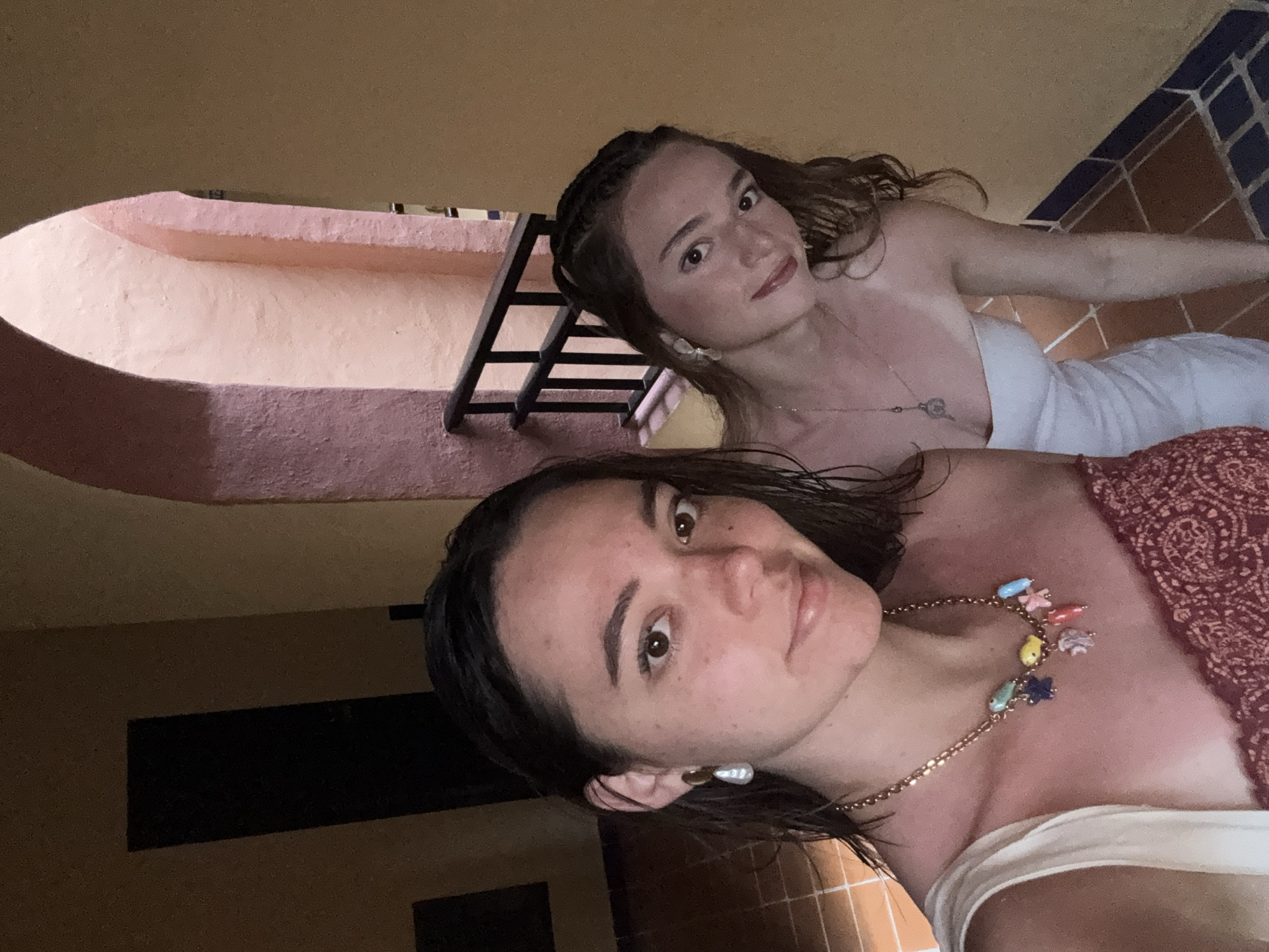 Two women are visible in an indoor setting. The woman in the foreground has short, dark hair and is wearing a gold necklace with colorful charms and large pearl earrings. She is wearing a white top, and the top part of a red patterned dress is seen on her right side. The second woman, positioned behind and to the left of the first, has light brown hair styled with braids and is wearing a white dress and a silver necklace. Behind them is a pink arched wall, a black metal railing, and tiled flooring in shades of orange and blue. Based on the characteristics of the architecture and the tiled flooring, the location appears to be in Playa del Carmen, Mexico.