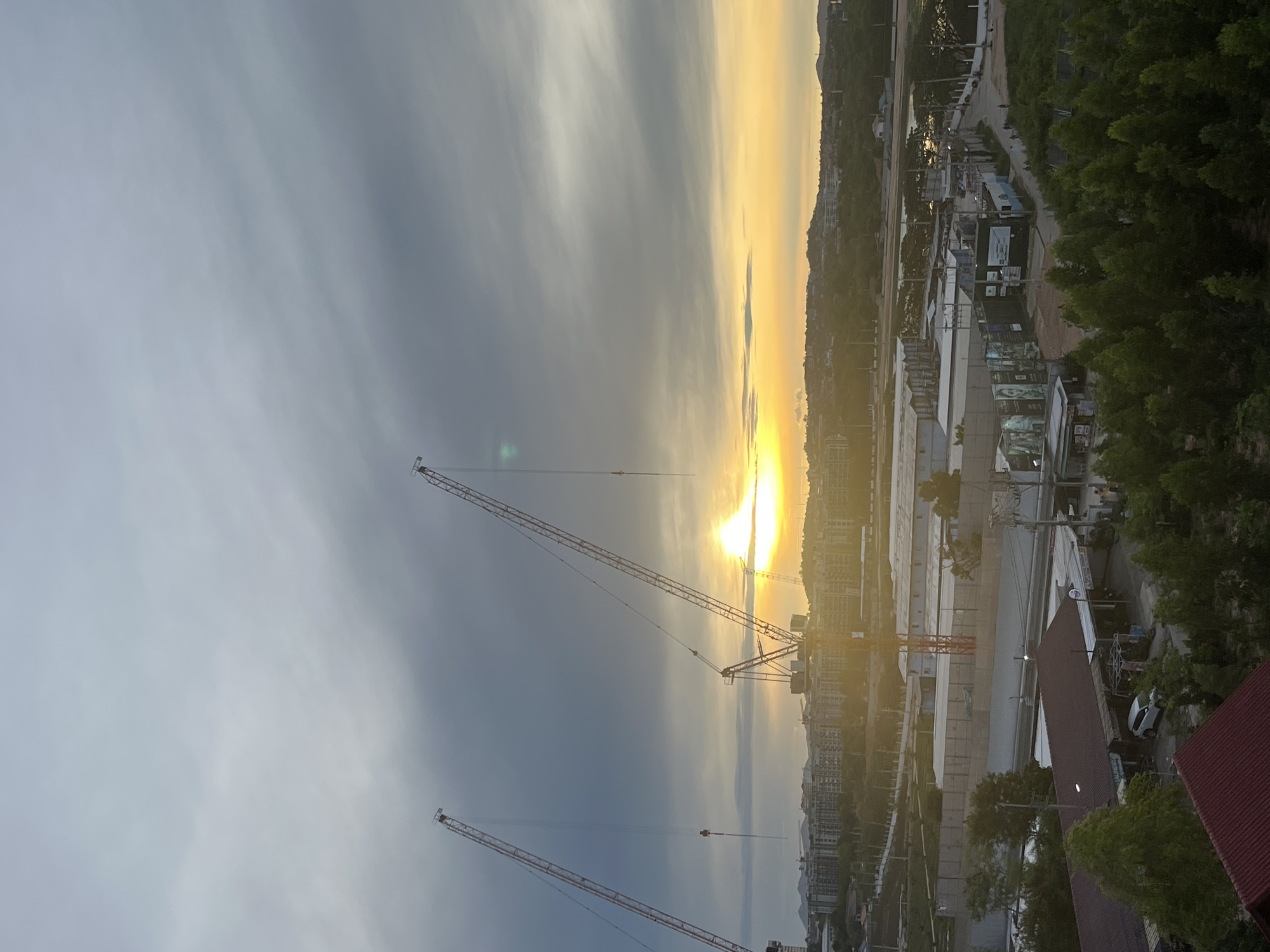 The image captures a cityscape in Pattaya City, Thailand, at sunrise or sunset. The sky is divided between gray clouds on the left and a bright, golden glow emanating from the horizon on the right. Several tall construction cranes are prominent in the foreground, their outlines sharp against the sky. Below the horizon, buildings and structures are visible, appearing somewhat hazy due to the lighting. Trees and vegetation are present on the far right, and part of a red rooftop can be seen in the lower right corner.