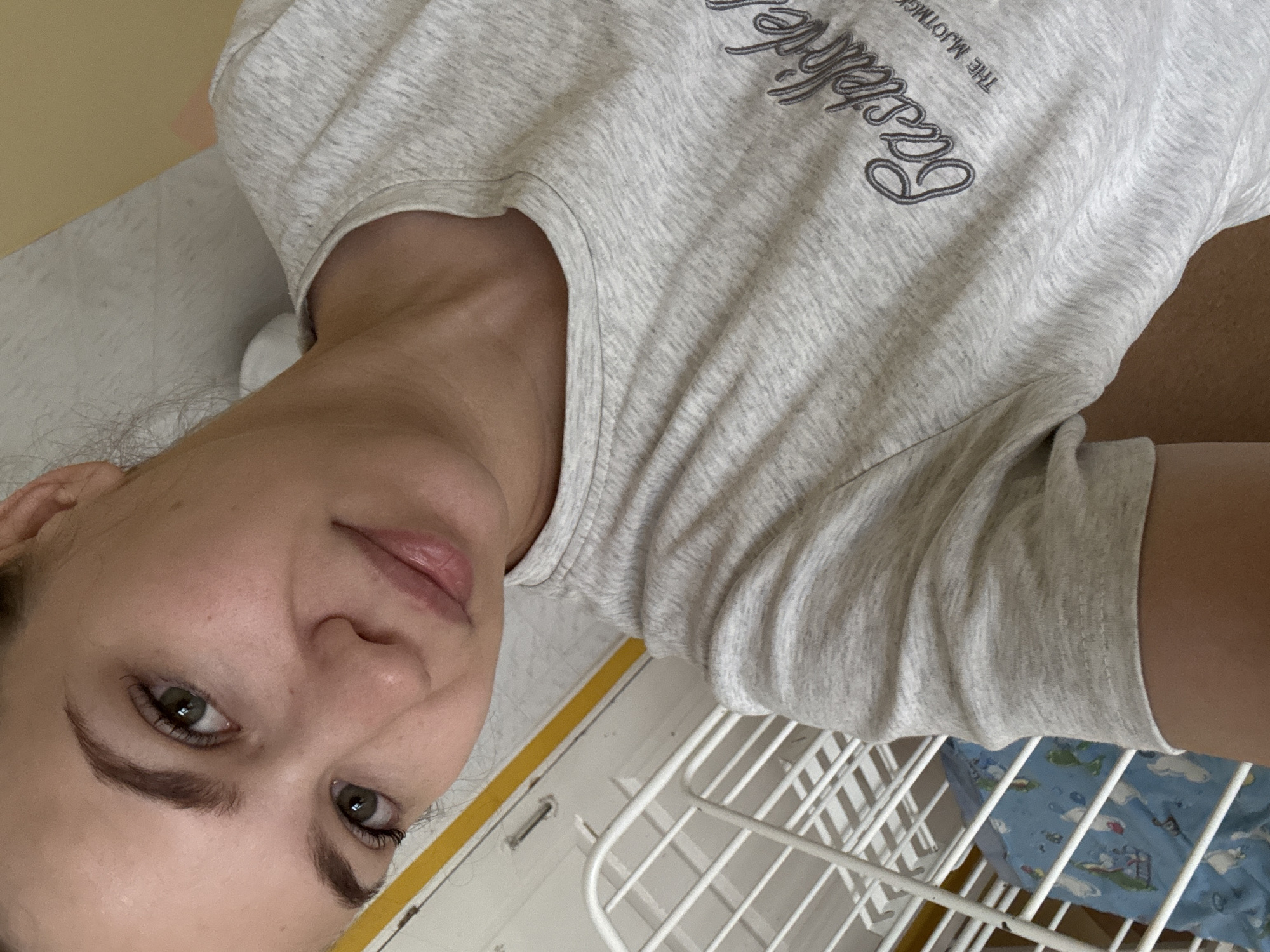 This image captures a close-up selfie of a young adult female, reportedly located in Chaikovsky, Russia. The woman has light skin, dark eyebrows, and hazel-green eyes that are open and directed towards the camera. Her dark hair appears to be pulled back, with a few lighter wisps visible near her left ear. Her lips are full and slightly parted, displaying a natural, soft pink hue. She is wearing a light gray heathered t-shirt with a rounded neckline. The front of the shirt features partial text, "Castelde" (or similar) in a script font, and below it, in a smaller sans-serif font, "THE MJOTMG," both in a darker gray or charcoal color. The perspective suggests she is either lying down or reclining, holding the camera above her.
The background indicates an indoor setting. To the left side of the frame, a white grated structure, resembling a baby crib or playpen, is partially visible. Inside this structure, a blue fabric with a light-colored map or continent print is discernible. Above this structure, a distinct horizontal yellow stripe runs across a white vertical surface, possibly a wall or door frame. To the upper right, a plain, light beige-colored wall or surface is visible, with a small portion of a white pillow or bedding below it.