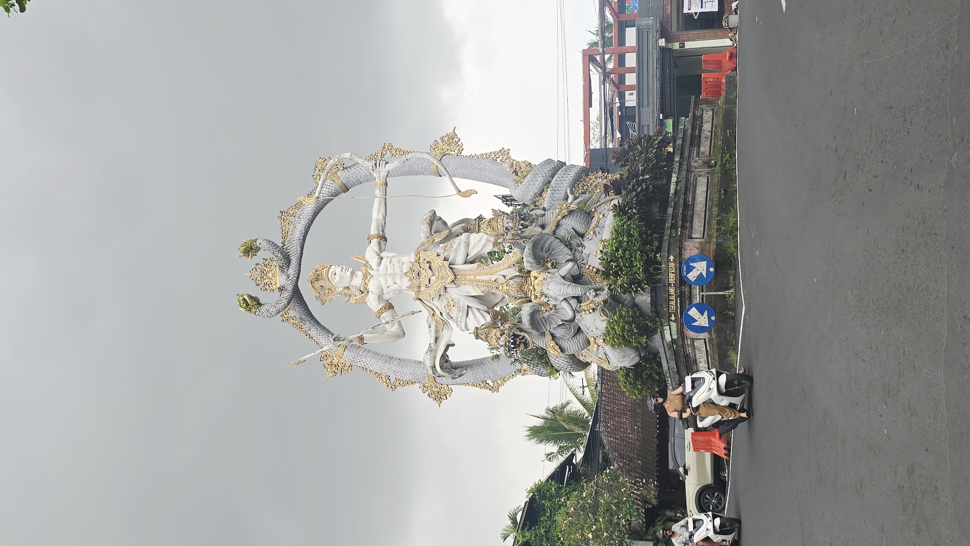 The image captures a vibrant street scene in the Ubud District, Indonesia. A prominent statue of a deity wielding a bow and arrow is surrounded by a multi-headed serpent. Traffic arrows are displayed on the roadside, and the road surface is visible with a person on a scooter driving to the left. Buildings and lush greenery define the background.