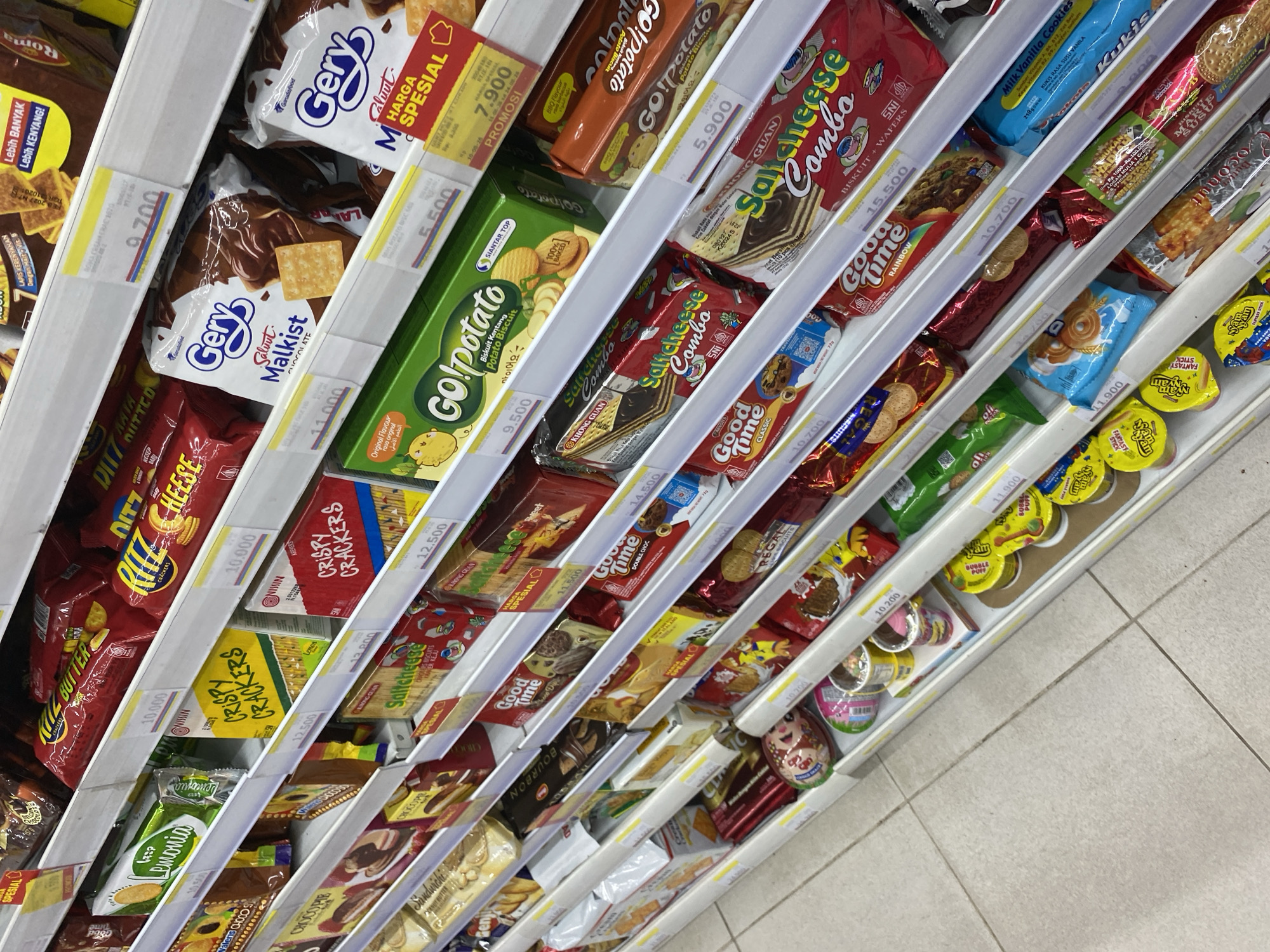 The image shows a retail shelf filled with various packaged snacks, primarily cookies and crackers, in a brightly lit store. The items are neatly arranged on white shelves. Several product brands are visible, including Ritz, Gery, Go! Potato, Good Time, Saltcheese, and MilkVanilla. Many of the packages have price tags or promotional labels with prices in Indonesian Rupiah. The overall environment suggests an aisle in a supermarket or convenience store.