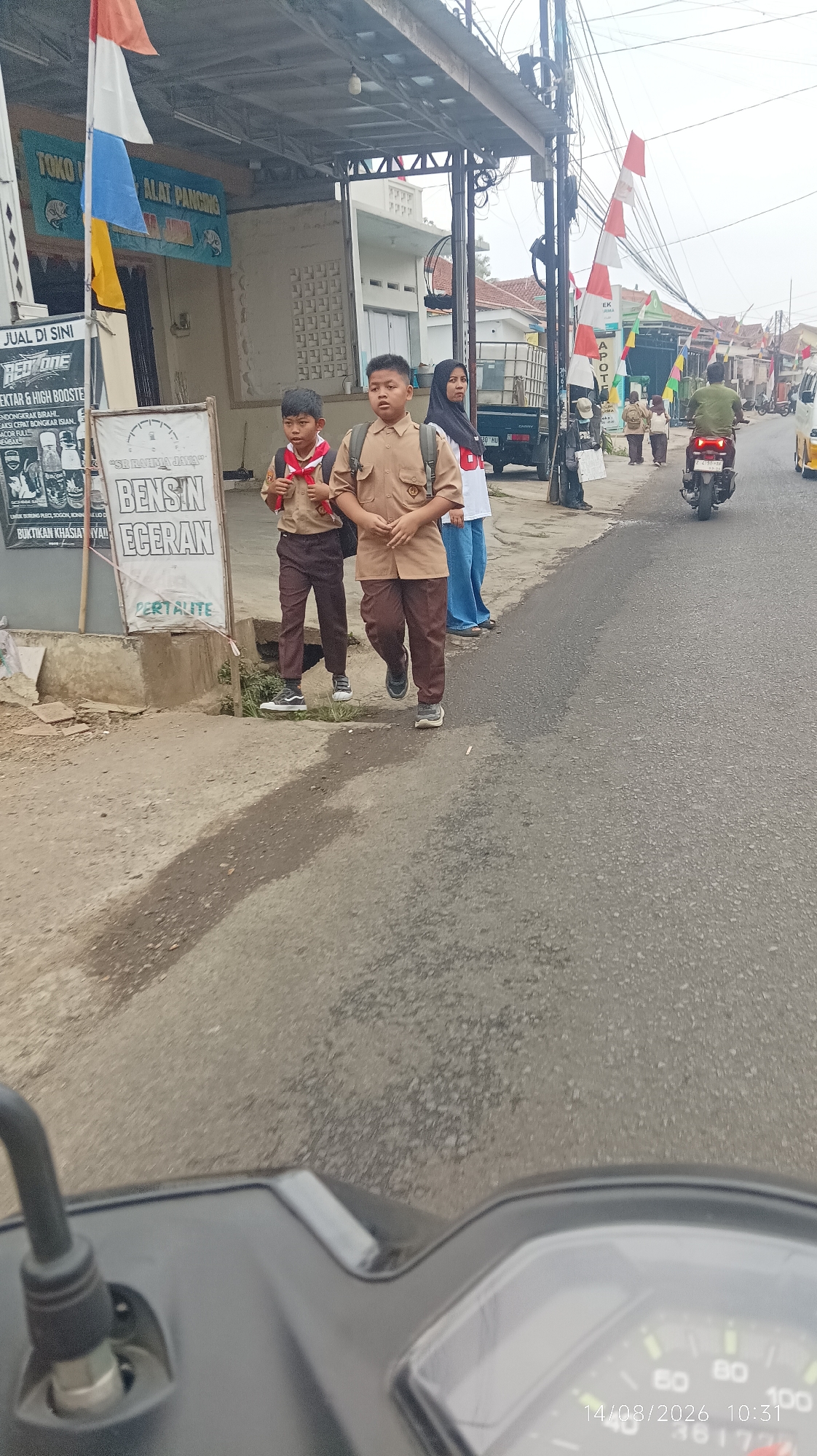 In Tasikmalaya, Indonesia, two young boys in tan school uniforms with red and white scarves walk on a sidewalk next to a street. The boy on the left is shorter than the boy on the right, and both wear backpacks. Behind them, a young woman in a white shirt and blue pants, and a black headscarf, walks with them. A man on a motorcycle rides on the street behind them, and several other people and motorcycles are visible further down the street. Shops line the left side of the street, with flags and advertisements displayed. A truck is parked in front of one of the shops. The sky is overcast, and the road is paved with asphalt. The image appears to be taken from the perspective of someone riding a motorcycle.