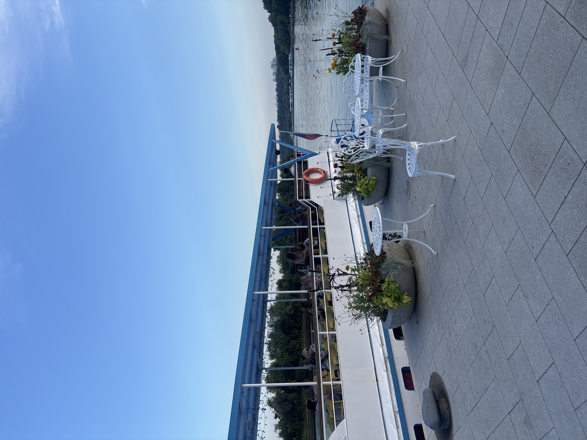 The image shows a waterfront scene in Moscow, Russia. A white tour boat with a blue canopy is docked along a paved promenade. Several people are seated on yellow chairs inside the boat, enjoying a view of the water. On the promenade, white wrought-iron tables and chairs are set up, some with decorative planters filled with flowers. The water is calm, reflecting the clear blue sky. Trees line the opposite bank of the river, and a distant cityscape is visible on the horizon. The atmosphere appears peaceful and leisurely.