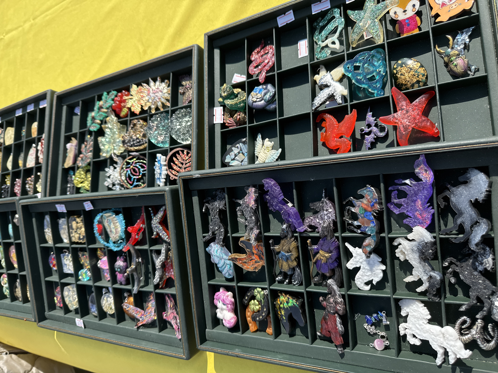 The image captures a vibrant display of handmade resin trinkets or brooches, presented in several dark green compartmentalized trays at what appears to be an outdoor market or craft fair in Chaikovsky, Russia. The bright sunlight, casting distinct shadows, suggests a clear and sunny day.
The primary subjects are the numerous unique artisanal pieces, each carefully arranged within its own section in the display cases. The trinkets are crafted from a translucent material, likely resin, often incorporating glitter, iridescent flakes, or colorful pigments, giving them a sparkling and ethereal quality. The designs are highly varied and imaginative, depicting a wide array of figures. These include fantastical creatures like dragons, unicorns, and various mythological beasts, alongside animals such as foxes, owls, snakes, horses, starfish, and beetles. Other designs feature stylized leaves, flowers, and abstract shapes, as well as human-like figures adorned in traditional garb, some carrying staffs. The color palette is rich and diverse, ranging from vibrant reds, purples, and blues to shimmering silvers, golds, and natural hues.
The display cases are neatly organized, highlighting each individual piece. Some of the items have small white price tags affixed, though the text on them is illegible. The backdrop to this colorful array is a bright, sunny yellow fabric, possibly a tablecloth or a tent awning, reinforcing the setting of an outdoor stall.
The overall scene depicts the orderly presentation of handcrafted goods for sale, indicating an event like a local market or festival where artisans showcase their work. The intricate details and diverse forms of the trinkets suggest a focus on unique, decorative items, potentially worn as jewelry or accessories.