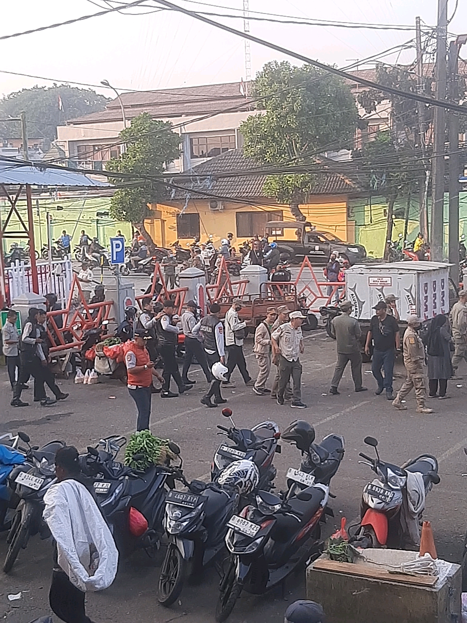 In the city of Bekasi, Indonesia, a street scene unfolds with a group of individuals, many in uniform, gathered around a street corner with parked motorcycles and temporary barricades. The environment suggests a public space, possibly near a market or transit hub, with several buildings visible in the background, one of which appears to be a school or administrative building with a prominent Indonesian flag.
The foreground is dominated by parked motorcycles, some of which have personal items like helmets and bags attached. A few individuals are seen interacting with each other, some appearing to be law enforcement officers in dark uniforms, possibly on duty, as indicated by their attire and the presence of barricades. One individual, wearing an orange vest, stands out amongst the others.
The middle ground shows more individuals, including some in what appear to be security or official uniforms, milling about near the barricades. There is a cart with produce, possibly being transported or sold. Further back, more motorcycles and a black pickup truck are visible, indicating vehicular activity.
The background reveals a cluster of buildings, some with tiled roofs and windows, and lush green trees. The overall atmosphere appears to be one of routine activity or perhaps a temporary situation being managed. The sky is bright, suggesting it is daytime.