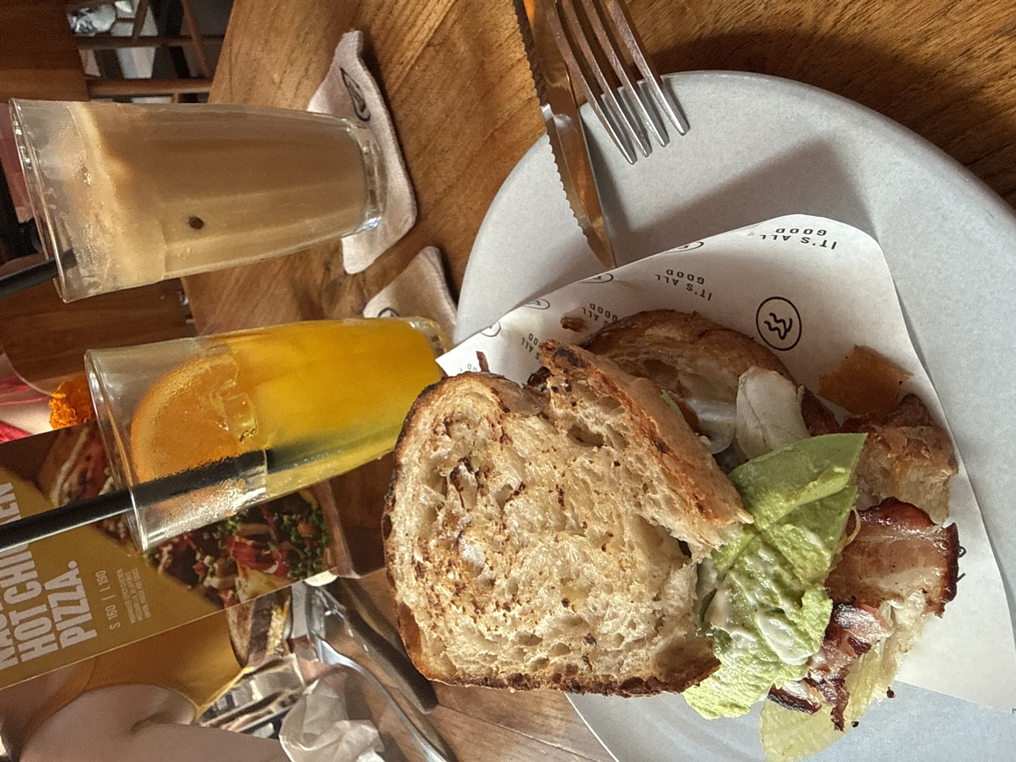 An overhead, slightly angled shot captures a meal laid out on a wooden table, likely in a cafe or restaurant. The central focus is a plate containing what appears to be a deconstructed sandwich or open-faced toast. It features a thick slice of grilled or toasted bread, accompanied by crispy bacon, and green elements resembling lettuce or avocado, with a creamy sauce. A piece of flaky pastry, possibly a croissant, is also visible. The food rests on a white paper liner with the repeating phrase "IT'S ALL GOOD" and a stylized wave-like logo printed on it.
Two beverages are present: one glass holds a light brown, frothy drink, likely an iced coffee or latte, with a single dark speck visible within the liquid. The other glass contains a vibrant orange liquid, presumably orange juice, garnished with an orange slice and featuring a black straw. A fork and knife are placed on a white napkin next to the plate. Part of a menu is visible on the table, displaying text such as "HOT CHI", "PIZZA.", "S 160 | L190", "MARNIER SPICY CHICKEN", and "MOZZARELLA PEPPER". The scene is well-lit, suggesting daytime. The overall impression is a casual dining experience in the Ubud District, Indonesia. No individuals are depicted.