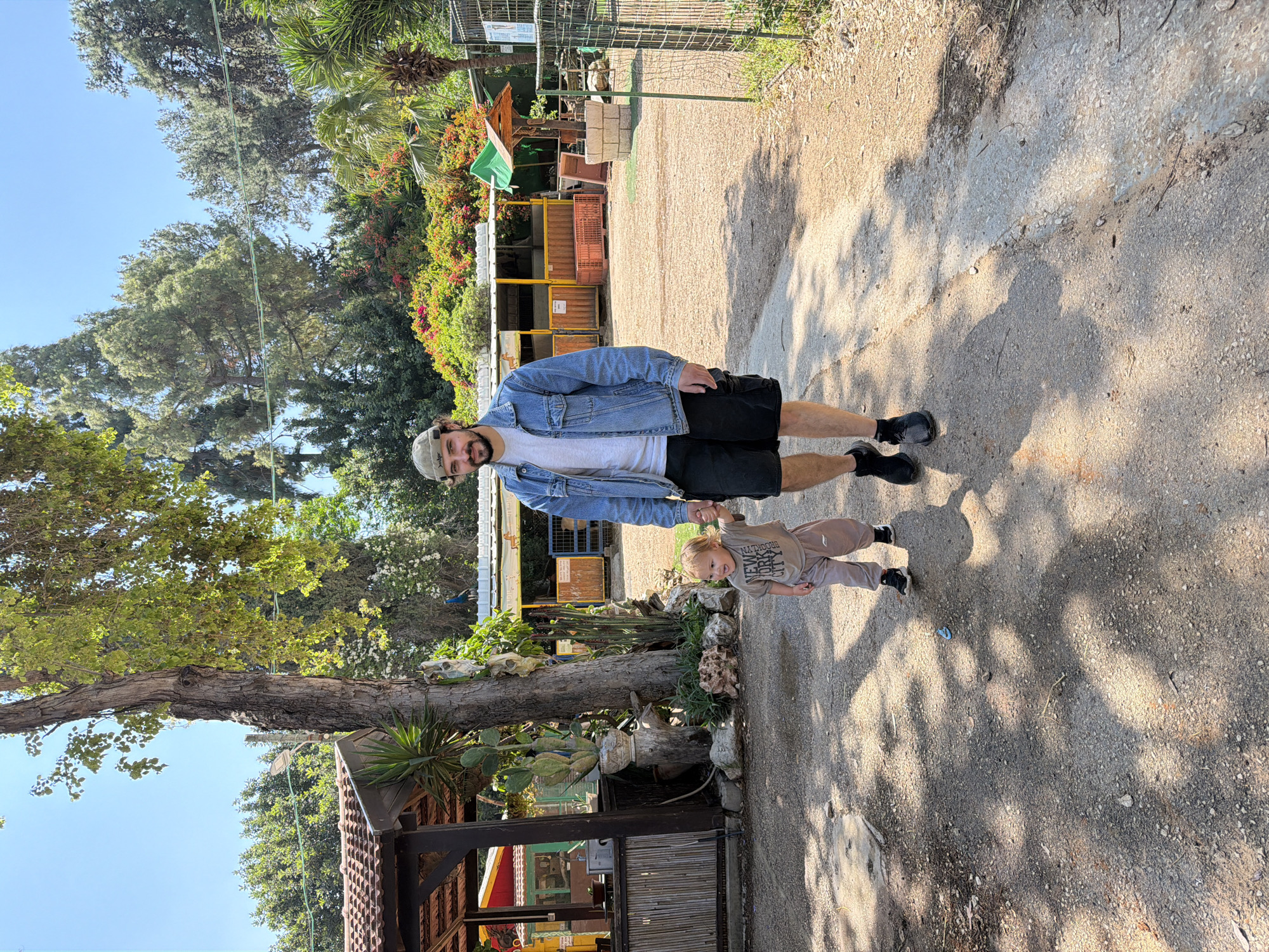 A man and a young child are walking together on a dirt path. The man is wearing a denim jacket, a white t-shirt, and black shorts. The child is wearing a brown shirt and pants. The background includes trees, a building, and fencing. The location is likely the city of Zvulun Regional Council, Israel.