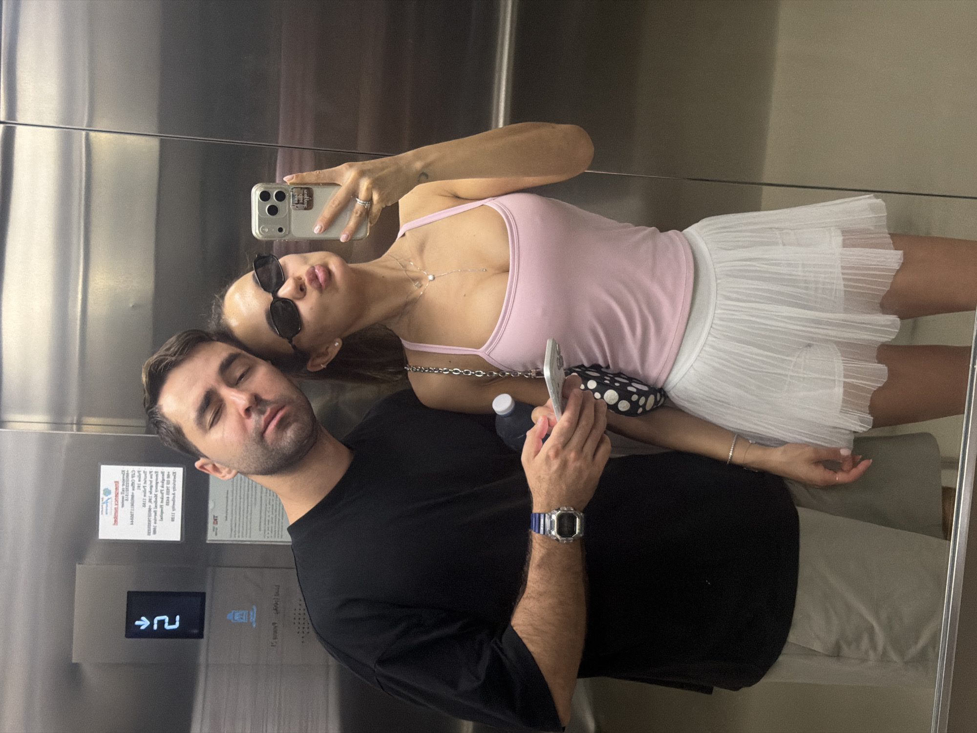A man and woman take a selfie in an elevator in Choeng Thale, Thailand. The woman, wearing sunglasses, a pink tank top, and a white skirt, holds a smartphone in her right hand to capture the image. She is leaning against the man who is wearing a black shirt and khaki shorts and stands beside her. The man appears to be sleeping, as he has his eyes closed. He has a blue watch on his left wrist. The elevator is silver and features informational posters and floor selection buttons. A water bottle is also visible next to the woman.