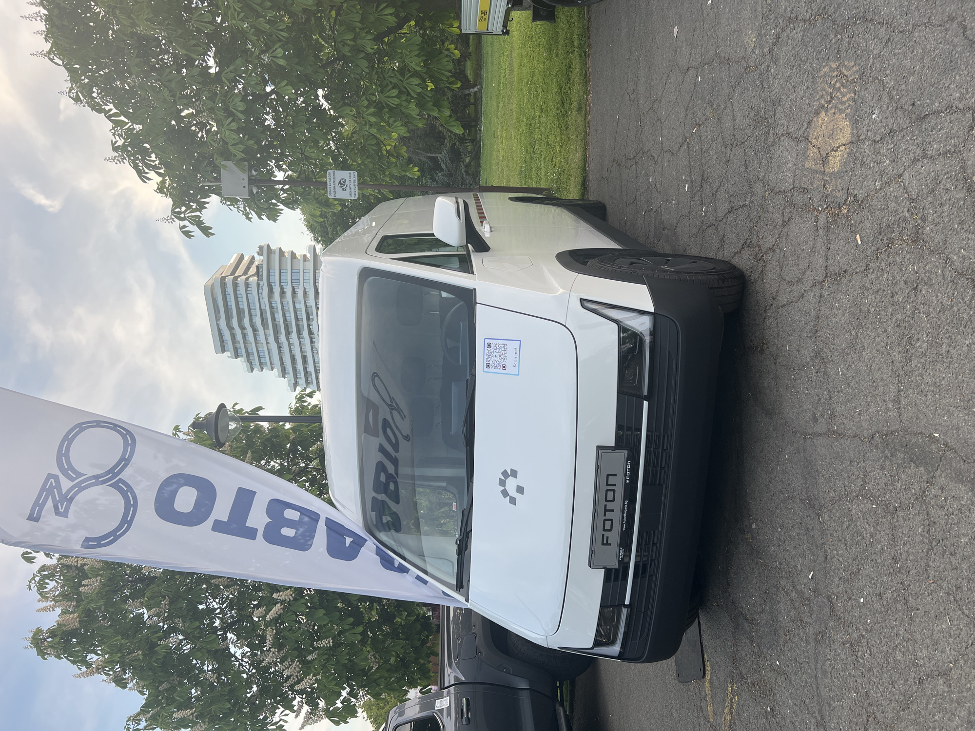 The image shows a white Foton brand electric vehicle parked on a paved surface, likely a parking lot or street. The front of the vehicle is prominently displayed, featuring the Foton logo and Bulgarian license plate. A white flag with blue lettering, possibly related to an automotive company or event, is positioned next to the vehicle.
In the background, there are green trees and grass, suggesting a park or landscaped area. A modern, multi-story building is also visible in the distance.
Text elements identified in the image include "ABTO", "FOTON", "www.Foton Bulgaria.bg," and "Зона с 24-часово AREA WITH 24 HOUR VIDEO SURVEILLANCE", indicating the presence of video surveillance. A QR code is also on the vehicle.