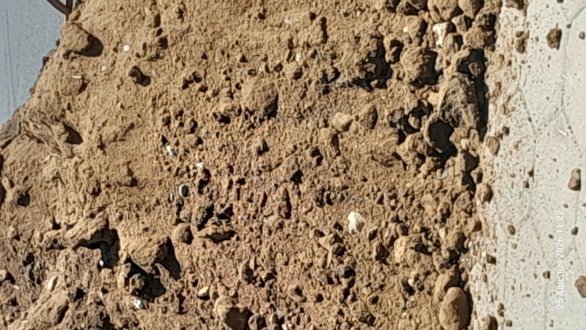 The image displays a close-up, angled view of a pile of dirt and rocks. The texture of the material is rough and uneven, with various sizes of stones mixed into the soil. Some areas appear compacted, while others show looser debris. In the lower part of the image, the dirt pile transitions to a lighter, smoother surface, possibly concrete or asphalt, scattered with smaller pebbles. A date stamp in the bottom left corner reads "25 Agustus 2026 11.22". The scene appears to be an outdoor environment, likely a construction or landscaping site, given the presence of dirt and debris. No people are visible in the image. The location is specified as Jomblang, Indonesia.