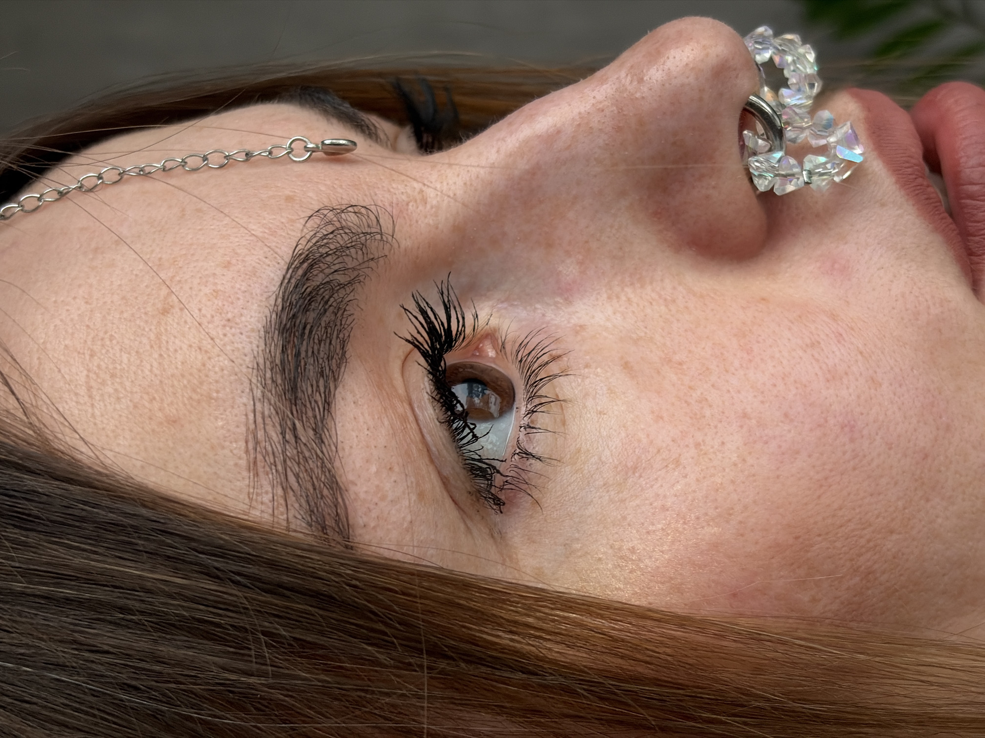 A close-up photograph depicts a woman's face in profile. The subject has fair skin with freckles and brown hair. She has long, dark eyelashes and a brown eye. A silver chain adorns her forehead, and a decorative nose ring with clear beads is visible on her nose. The background is blurred. The location is not discernible from the image.