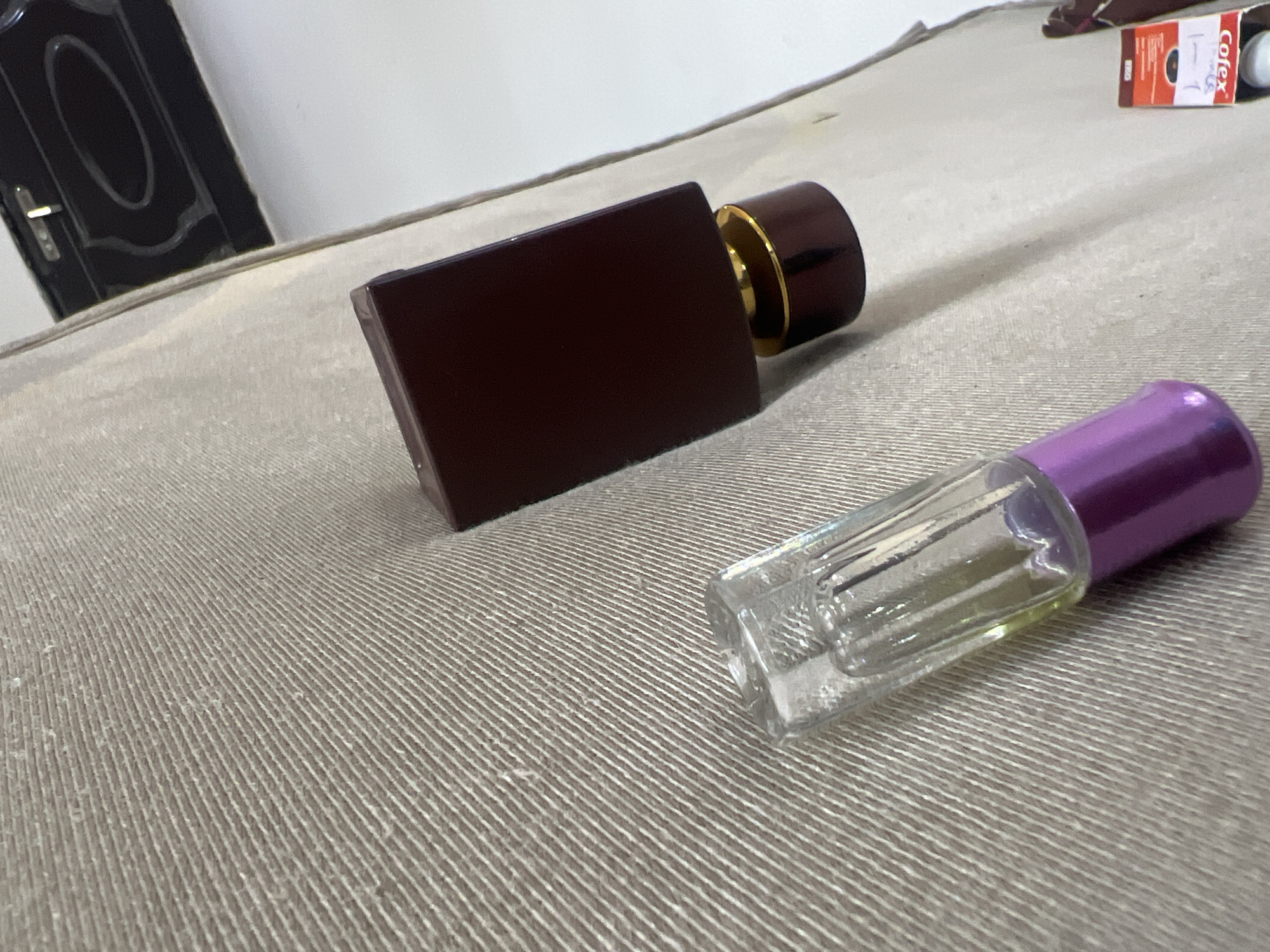 The image shows two perfume bottles on a light-gray textured surface. One bottle is a dark reddish-brown rectangular shape with a gold-colored neck and a dark brown cap. The other bottle is clear with a purple cap and a partially visible nozzle inside. In the upper right corner, a box labeled "Cofex" is visible. The background is blurry, but a dark brown door is discernible on the left side of the frame. It is not possible to identify the location as Abuja, Nigeria or any other specific location based on this information.
