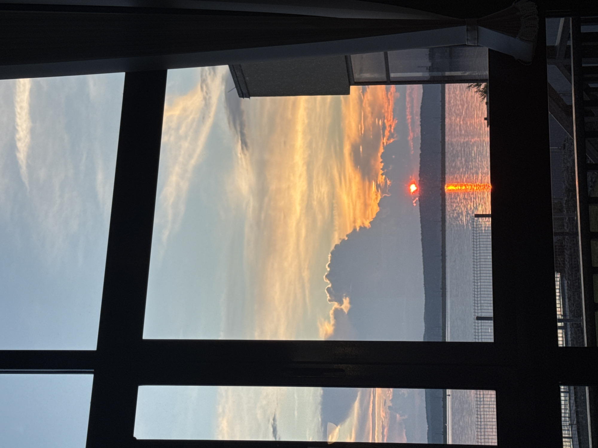 The image depicts a sunset viewed through a large window frame with dark mullions. The sky is filled with dramatic clouds painted in hues of orange, yellow, and pink, with streaks of lighter blue. The sun, a vibrant red disc, is partially obscured by a large cumulus cloud and casts a shimmering, golden reflection on the calm water below. A distant, dark, tree-lined horizon separates the water from the sky. In the foreground, through the window, parts of a building with a textured facade are visible, along with a white curtain. The view suggests a location overlooking a body of water, possibly the Kama River, in Chaikovsky, Russia, given the characteristic landscape of the region.