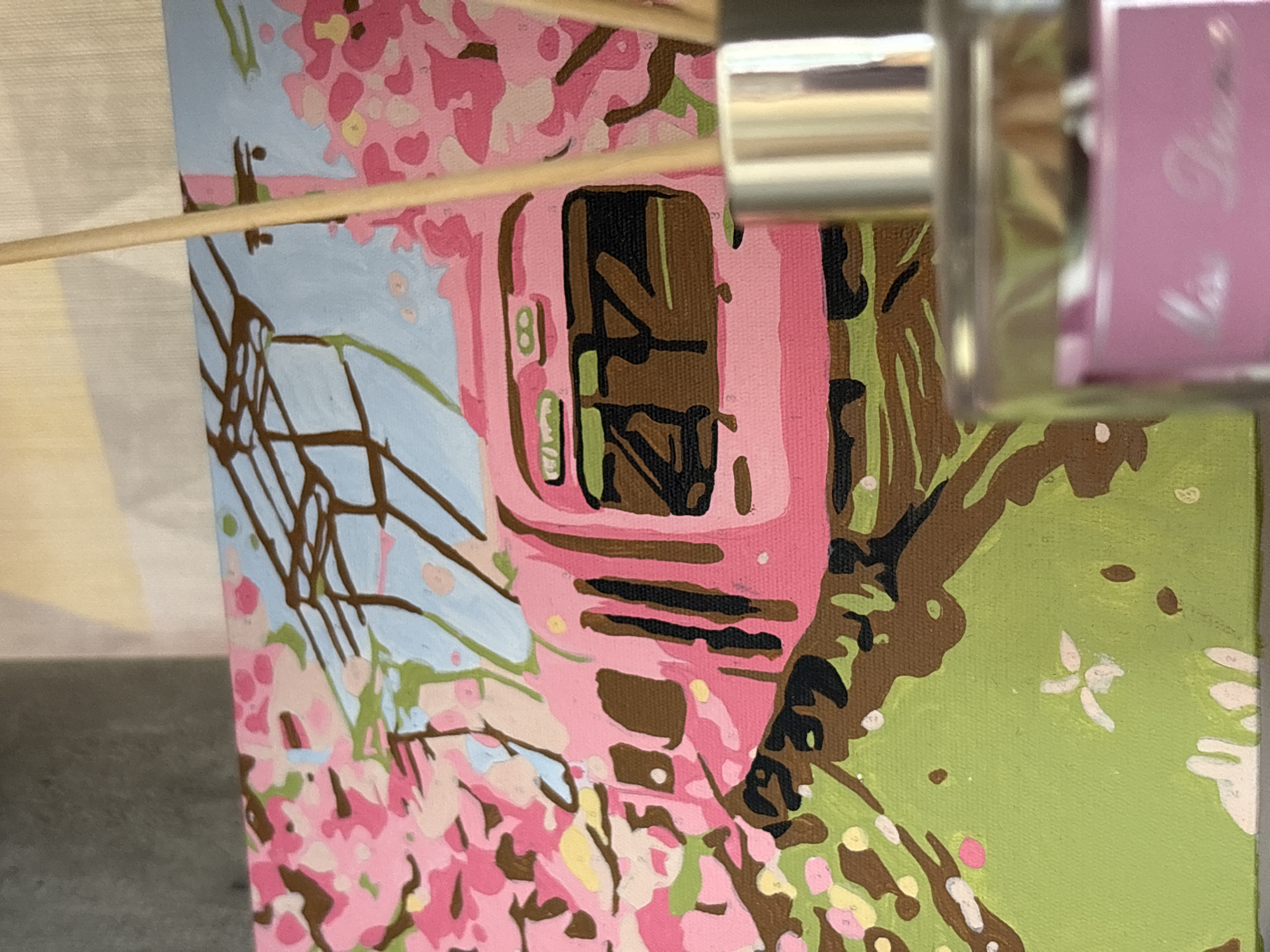 A close-up shot shows a partially completed paint-by-numbers canvas. The subject appears to be a building with pink exterior walls and a dark brown rectangular window. The background features a light blue area and trees with pink blossoms. A paintbrush rests horizontally across the top portion of the canvas. The right side of the frame is dominated by blurred bottles, one of which is pink. The painting is likely taking place indoors. The location mentioned is not visually identifiable within the scene.