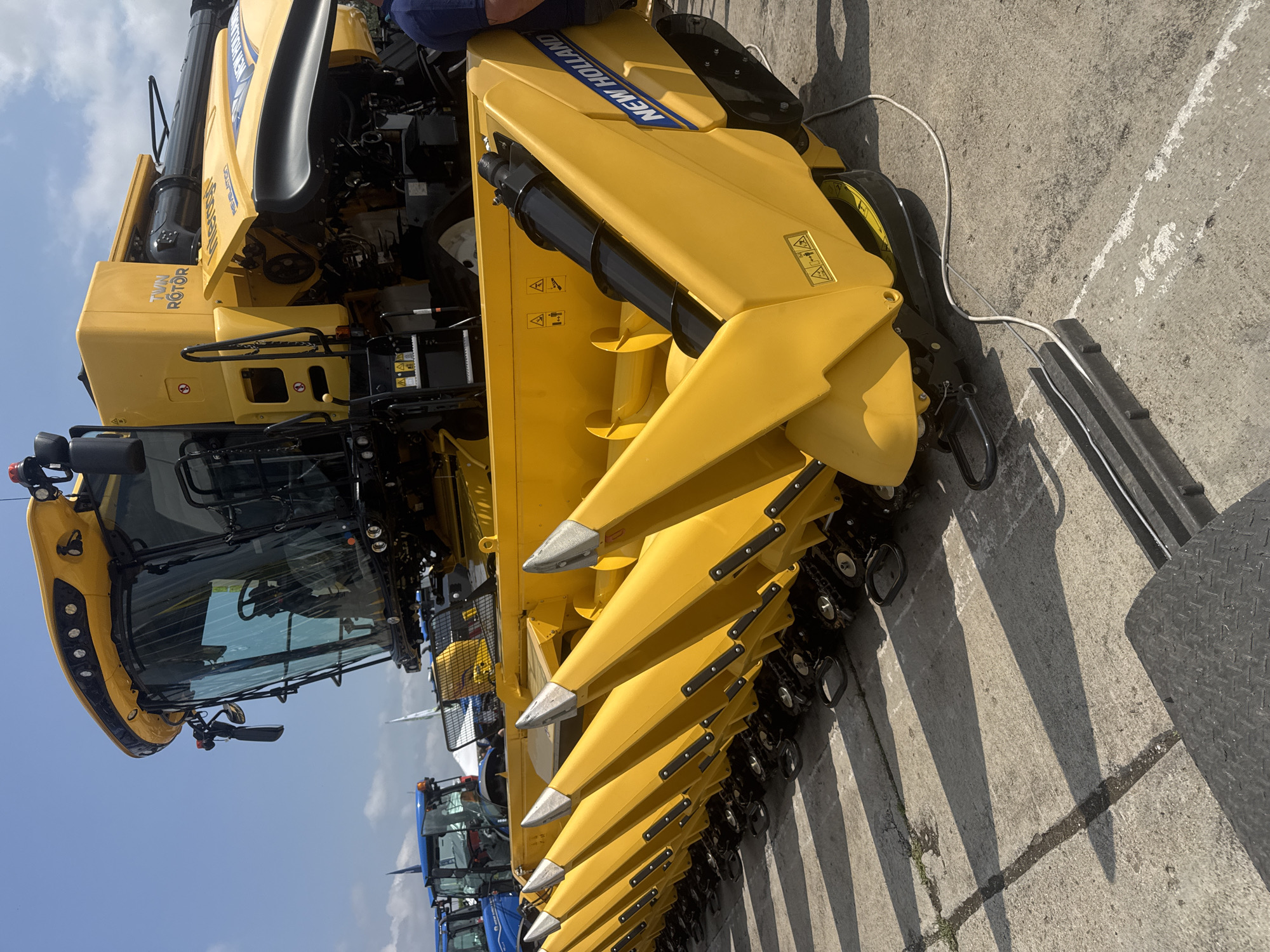 A large, bright yellow New Holland combine harvester is prominently displayed on a concrete surface under a clear blue sky in Stara Zagora, Bulgaria. The image, shot from a slightly elevated side angle, highlights the impressive scale of the agricultural machinery.
The combine features a "NEW HOLLAND" logo in blue letters on its side and "TWIN ROTOR" text, indicating a specific harvesting technology. Its front is equipped with a multi-row header, likely for harvesting corn, characterized by numerous pointed, yellow snouts with silver tips, designed to gather crops. These snouts cast long, sharp shadows on the ground, suggesting bright daylight, possibly morning or late afternoon.
A spacious cabin with large glass windows is visible on the left, offering a glimpse of a steering wheel inside. Partial views of other blue agricultural machinery, possibly tractors, are seen in the background behind the combine's cab. A white cable runs across the concrete ground near the combine's front right wheel, leading to a dark, textured mat or ramp.
The machinery appears to be stationary, likely parked for display at an event or dealership, rather than actively engaged in harvesting. Several warning and instructional pictograms, including triangular exclamation marks, are visible on various yellow panels of the combine. The overall scene conveys a sunny, outdoor setting, emphasizing the robust design of modern farming equipment.