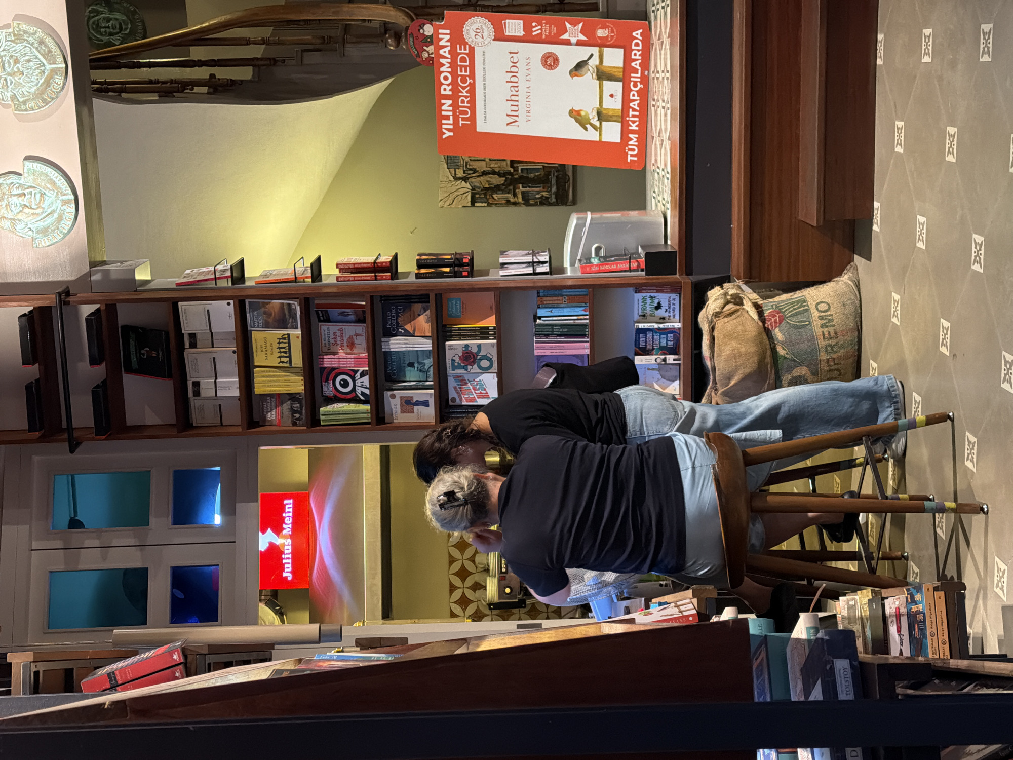 The image depicts the interior of a bookstore in Üsküdar, Turkey. Two women are seated at a counter, one with blonde hair wearing a black shirt and light blue jeans, and the other with dark hair wearing a black shirt and jeans. A wooden stool is positioned in front of the blonde-haired woman. To the left, a display of books is visible, including a large red sign that reads "Julius Meinl" in white lettering. Shelves filled with books line the walls, with a variety of titles in different languages. A staircase with a wooden railing is visible in the background. In the foreground, a large burlap sack with a floral design is placed on the floor. The floor is tiled with a pattern of white and grey geometric shapes. A promotional poster for a book titled "Muhabbet" is displayed on an orange stand. The overall impression is of a cozy and well-stocked bookstore, likely a place for reading and browsing.