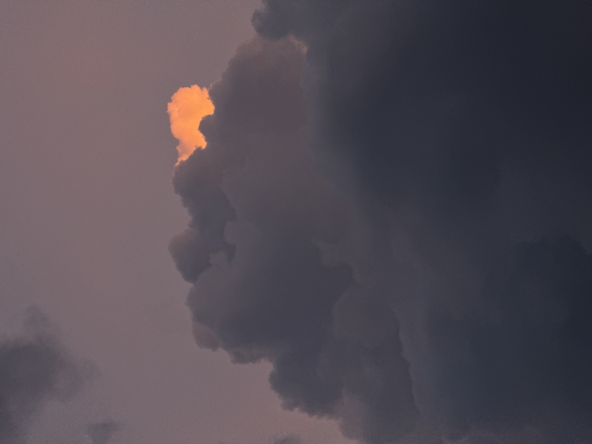 This is a close-up, vertical photograph of dark, moody clouds against a dusky purple sky. A small patch of cloud near the top center is brilliantly illuminated by a warm, orange glow, suggesting the sun is setting or has just risen behind it. The rest of the clouds are rendered in deep grays and purples, creating a dramatic and atmospheric effect. The image is devoid of any human elements, structures, or any indications of a specific location within Savenki, Russia. The focus is solely on the celestial phenomenon.