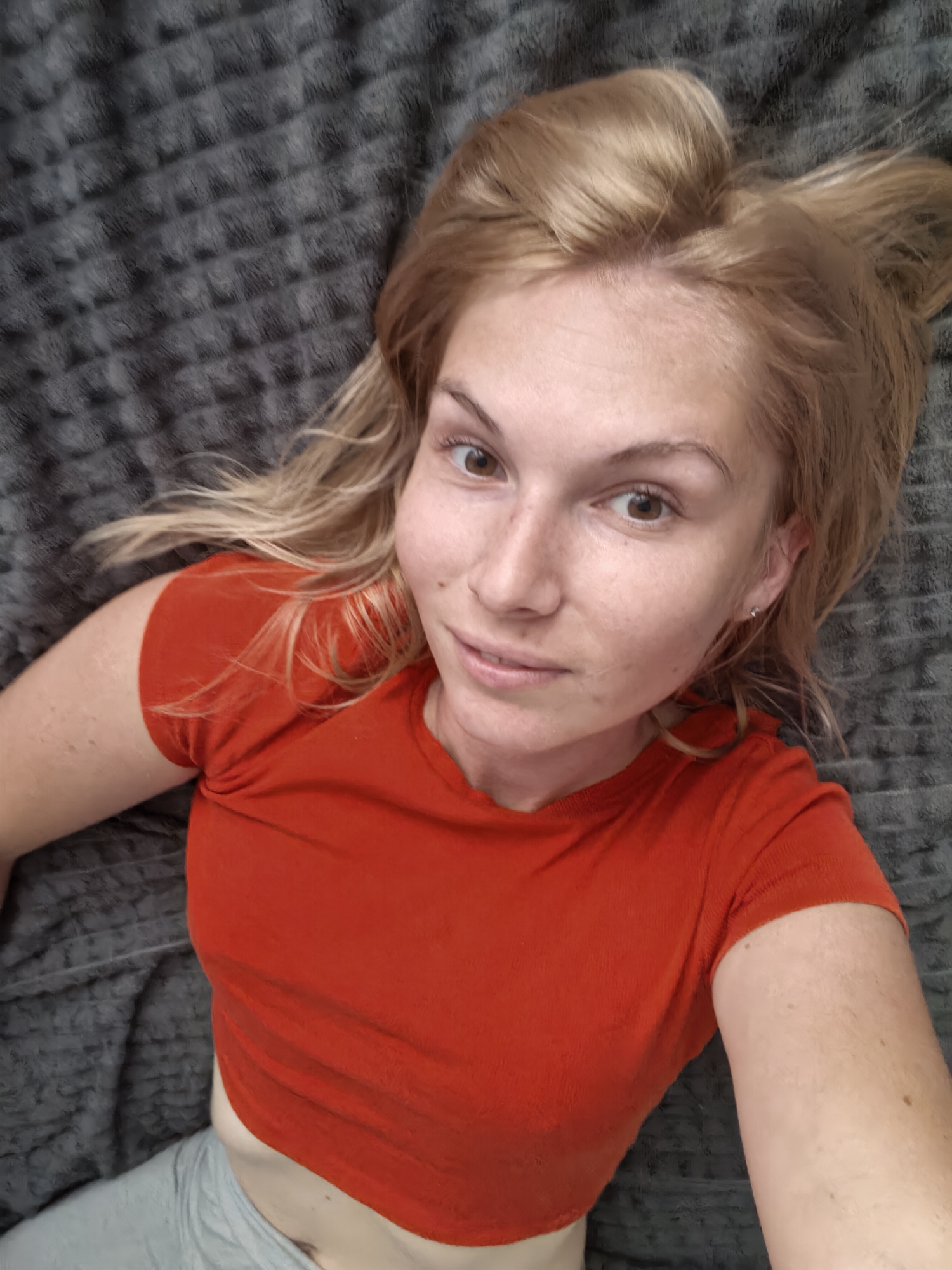 A blonde woman with light skin and freckles lies on a grey textured surface, looking up at the camera. She has brown eyes and is wearing a bright orange t-shirt and light grey pants. Her hair is light blonde and slightly messy. The image is taken from a high angle, looking down at the woman. There is no indication of the city of Sterlitamak, Russia, in the image.