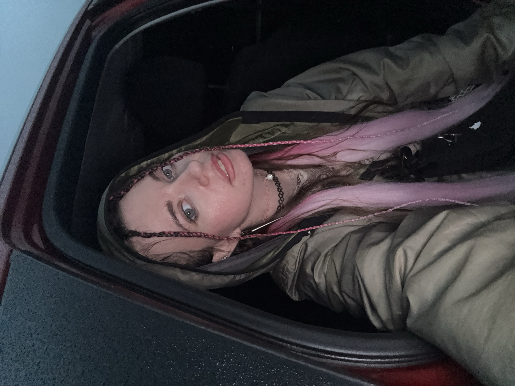 A young woman is pictured from the chest up, inside a vehicle, looking directly at the camera. She has light skin, blue eyes, and her hair is styled in braids with pink extensions. She is wearing a dark choker necklace with a pendant and a hooded, olive-colored jacket. The background is dark, suggesting it is either nighttime or the interior of the vehicle is dimly lit. The exterior of the vehicle is visible on the left side of the image, and it appears to be raining, with water droplets on the window. There is no identifiable location or context provided in the image.