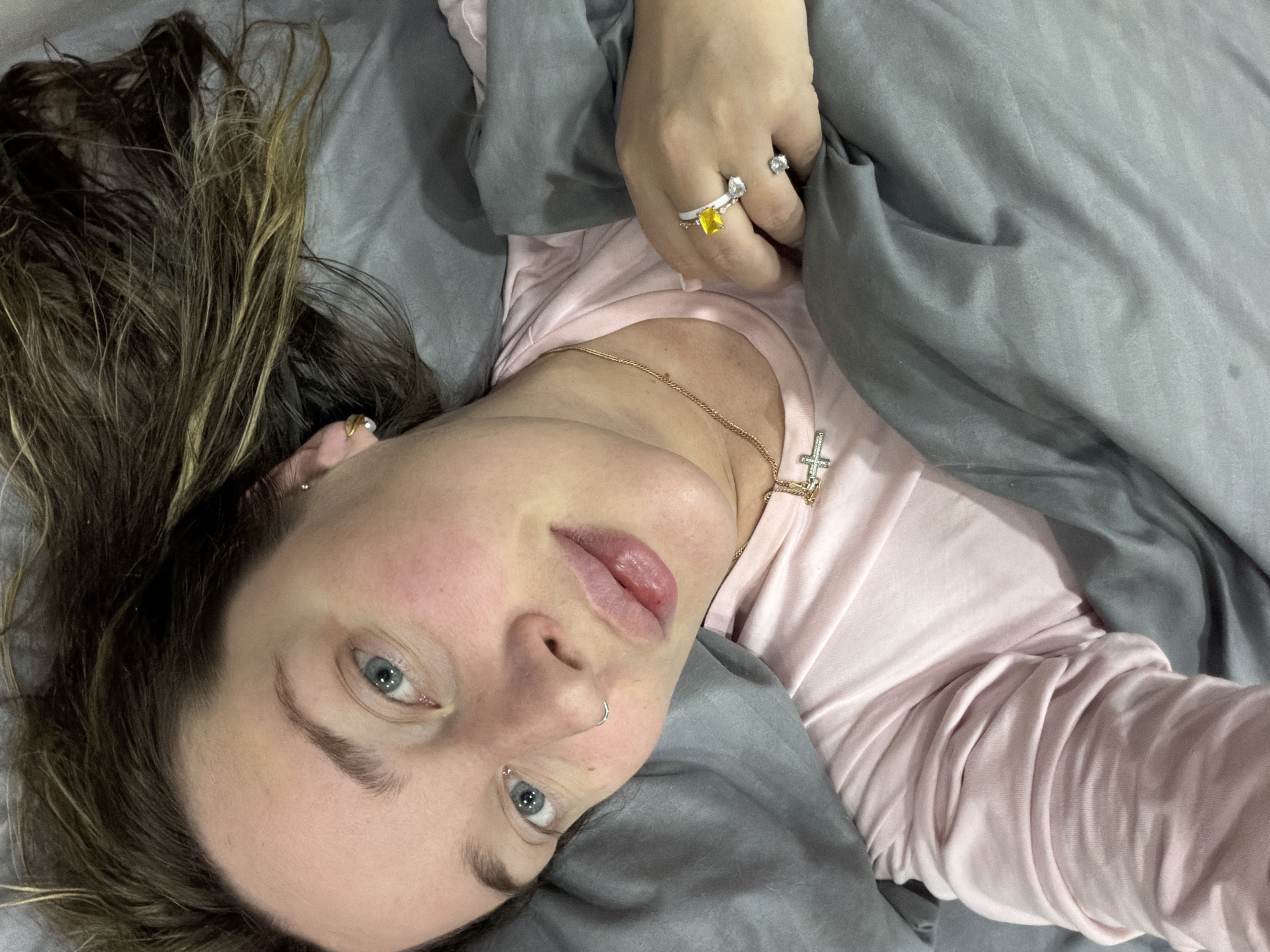 The image presents a close-up, high-angle portrait of a young woman lying in bed, likely in Chaikovsky, Russia. She is looking directly at the camera with a neutral, slightly relaxed expression, her blue eyes wide open. Her long, brown hair with lighter, possibly blonde, streaks is fanned out around her head on grey bedsheets, appearing somewhat disheveled, suggesting she might be waking up or relaxing.
She is wearing a light pink long-sleeved top. Adorning her are several pieces of jewelry: a delicate gold chain necklace with a small cross pendant, and on her right hand, multiple rings including one with a distinctive square yellow gemstone, another with a clear or white stone, and a plain band. She also has a small silver hoop nose ring in her left nostril and a subtle stud earring in her left earlobe. Her skin is fair, with a natural flush visible on her cheeks and slightly parted lips. The surrounding bedding is a soft grey color. The lighting is soft and even, indicating an indoor setting without direct harsh light. No text is visible in the image.
