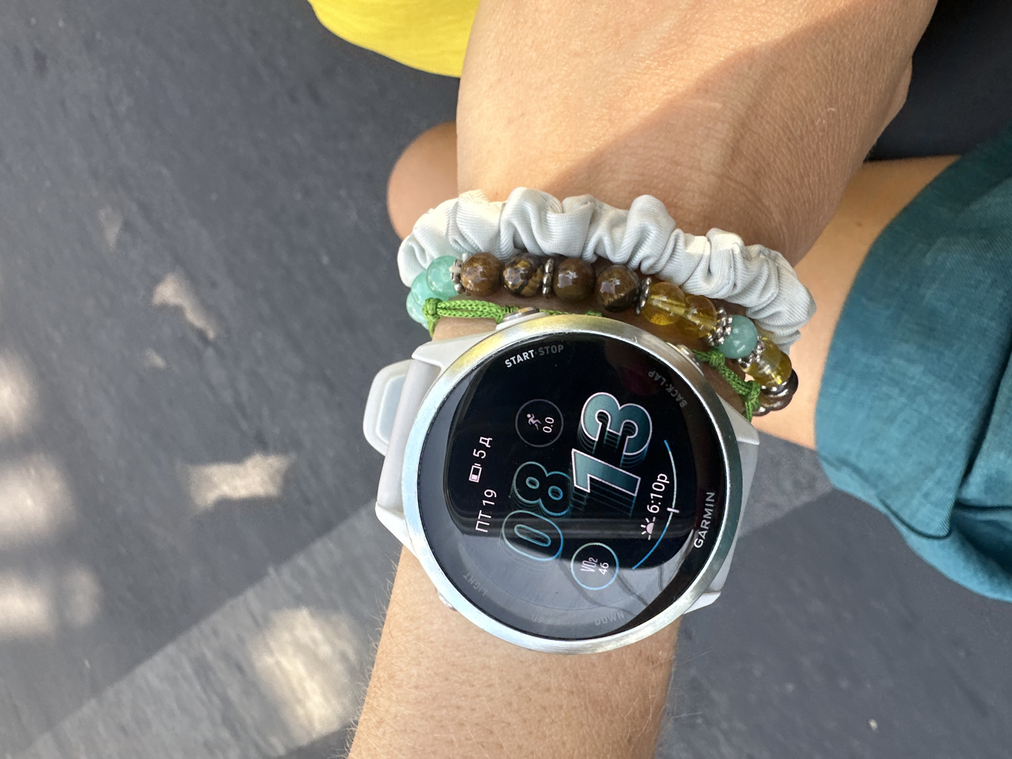 The image displays a close-up view of a person's wrist, adorned with a white Garmin smartwatch and several beaded bracelets. The smartwatch face shows the time "08:13", a date "ПТ 19" (likely Friday the 19th), a VO2 max reading of "46", and other activity metrics. The bracelets consist of brown and green beads. The background is a dark, textured surface, possibly asphalt, with some lighter, out-of-focus shapes that could be reflections or debris. The lighting suggests an outdoor setting, likely during the day, casting shadows. The image does not contain any contextual information that would allow for the identification of a specific location in Bac Nha Trang Ward, Vietnam.