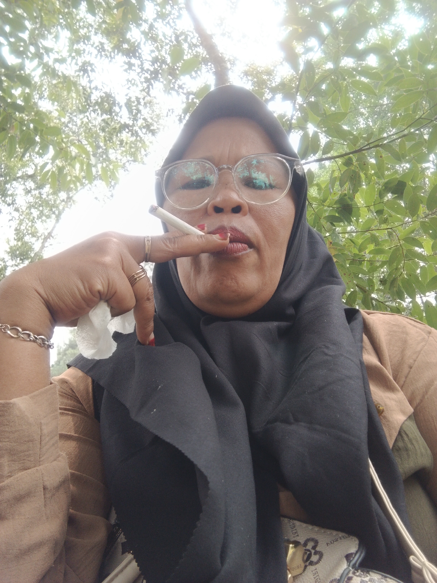 An outdoor, low-angle, medium shot shows a woman wearing a black hijab, glasses, and a brown top, holding a lit cigarette in her right hand. Her left hand is holding a crumpled white tissue. She has red lipstick on and jewelry on her fingers and wrist. The background consists of green foliage and a bright sky, suggesting a natural outdoor setting. The woman appears to be smoking the cigarette. The image was taken in Perbawati, Indonesia.