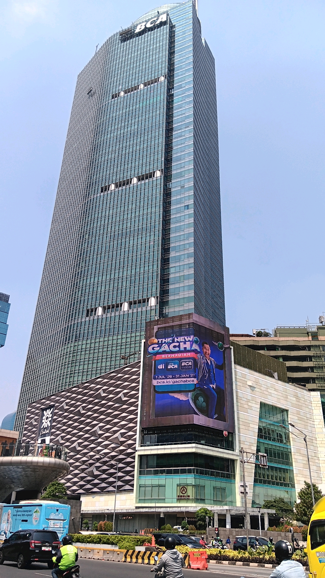 This is a low-angle exterior shot of the BCA Tower building in Central Jakarta, Indonesia. The skyscraper is clad in blue-tinted glass and features a prominent "BCA" logo at its apex. On the lower portion of the building, a large digital billboard displays an advertisement for "THE NEW GACHA" featuring a smiling man in a suit and text including "BERHADIAH" and "di BCA mobile." Below the billboard, a patterned facade with a zig-zag design is visible. In the foreground, a street scene unfolds with various vehicles, including cars, motorcycles with riders wearing helmets and vests, and a blue bus. The environment suggests a busy urban setting with a clear sky overhead.