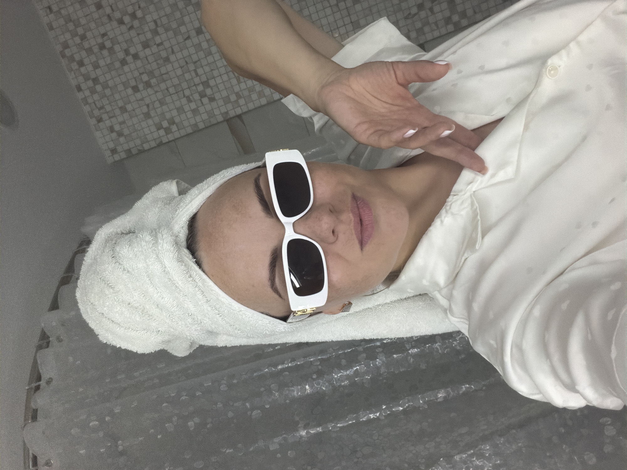 A woman lies on a surface covered by a semi-transparent textured material, likely a shower curtain, wearing white-framed sunglasses and a towel wrapped around her head. She is dressed in a light-colored, possibly silk, shirt. Her right arm is raised, with her hand gesturing near her chest. The background includes a wall tiled with small, square tiles. The location is a bathroom in Votkinsk, Russia.