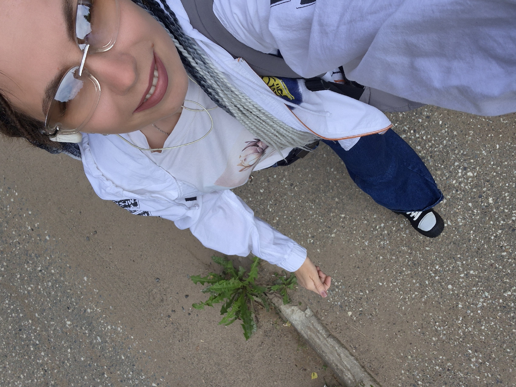 A young woman with braided hair and sunglasses is standing on a sidewalk in the city of Chaikovsky, Russia. She is wearing a white jacket, a white t-shirt with a floral design, and blue jeans. She is also wearing white headphones and a silver necklace. Her left hand is extended towards a plant growing through a crack in the sidewalk. The background shows a concrete sidewalk and some green vegetation. The image is taken from a high angle, looking down at the woman.